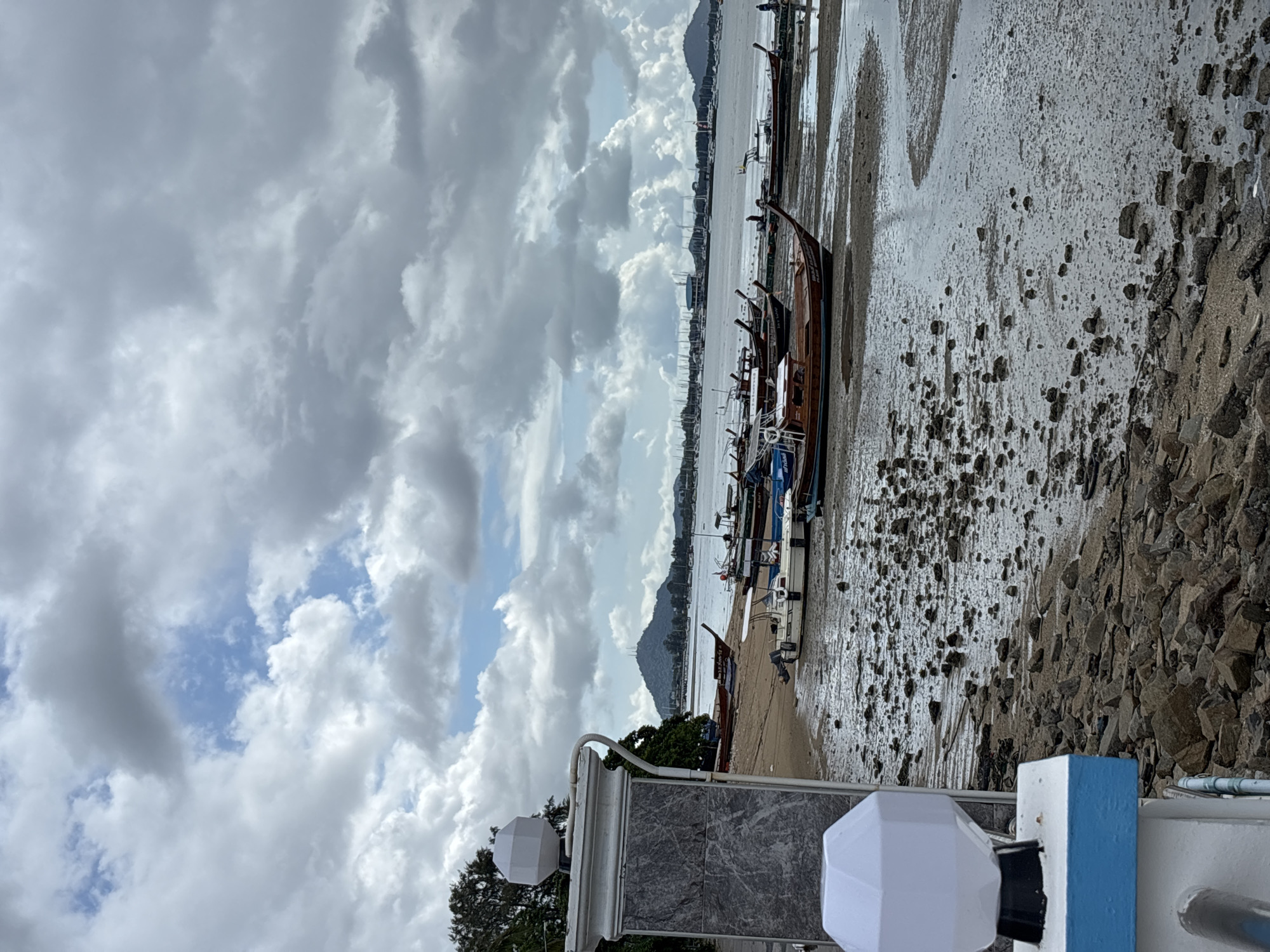 The image depicts a waterfront scene in Rawai, Thailand, characterized by a muddy shore dotted with numerous boats. The sky is filled with dramatic, cloudy formations, suggesting overcast weather. In the foreground, a modern white outdoor light fixture is mounted on a pillar adorned with gray stone-like material. Beyond the light fixture, a path or walkway leads towards the shore.
The shore itself is a mixture of wet mud and scattered rocks. Several longtail boats, a common type of vessel in Thailand, are moored on the beach or in shallow water. Some are wooden, displaying their characteristic slender hulls and elevated sterns. A few smaller motorboats are also visible, including one white boat with an outboard motor. In the distance, the water stretches out to a hazy horizon, with more boats and structures, possibly a marina or other waterfront development, visible. A lush green tree line runs along the left side of the image. The overall impression is one of a coastal area at low tide, with boats resting on the exposed seabed. A single person can be seen walking in the distance near the water.