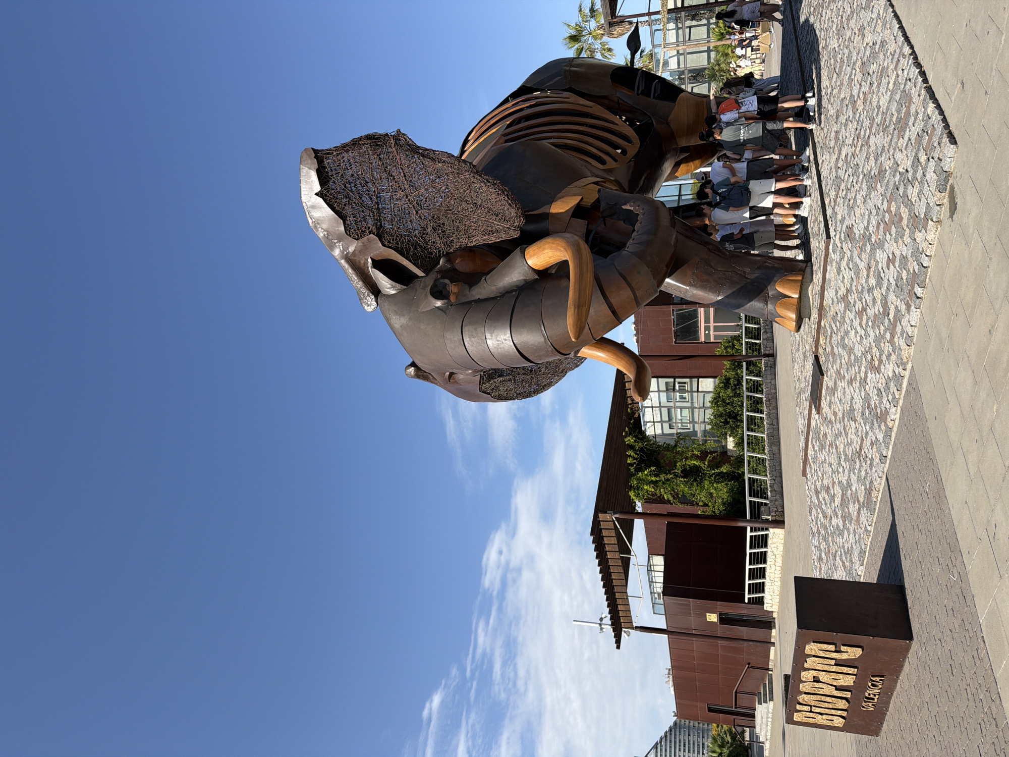 This vertical photograph shows a large, metallic sculpture of an animal, possibly a dinosaur or a prehistoric creature, on display outdoors. The sculpture is dark brown and appears to be made of metal, with visible internal framing and some woven wire elements. It is positioned in the foreground, leaning forward and slightly tilted.
Behind the sculpture, a group of people are standing in a line, looking towards the sculpture or away from the camera. They are dressed in casual summer clothing, with some wearing shorts and t-shirts. The setting is an urban outdoor space with a paved walkway made of small, irregularly shaped stones. Further in the background, there is modern architecture, including a brown building with large windows and some greenery. Palm trees are visible above the building. The sky is a clear, bright blue with a few wispy white clouds, suggesting it is daytime and the weather is fair.
In the lower right corner of the image, a dark brown wooden sign is placed on the ground. The sign has raised, metallic letters that spell out "BIOPARC VALENCIA". The overall impression is of a tourist attraction or an outdoor exhibition on a sunny day.