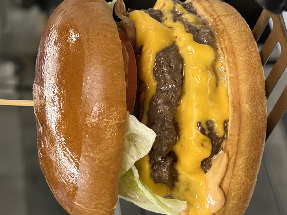A detailed, close-up image displays a double cheeseburger in Budva, Montenegro, presented on a metallic slotted spatula. The burger consists of a glossy, toasted brioche bun, secured vertically by a light-colored wooden skewer. Between the buns, from top to bottom, are layers of crisp light green lettuce, red tomato slices, and dark bacon strips. Two beef patties are stacked, each covered with melted bright yellow cheese that cascades over the edges. A creamy, light orange-pink sauce is visible on the bottom bun. The background is dark and out of focus, indicating an indoor setting. No individuals are visible.