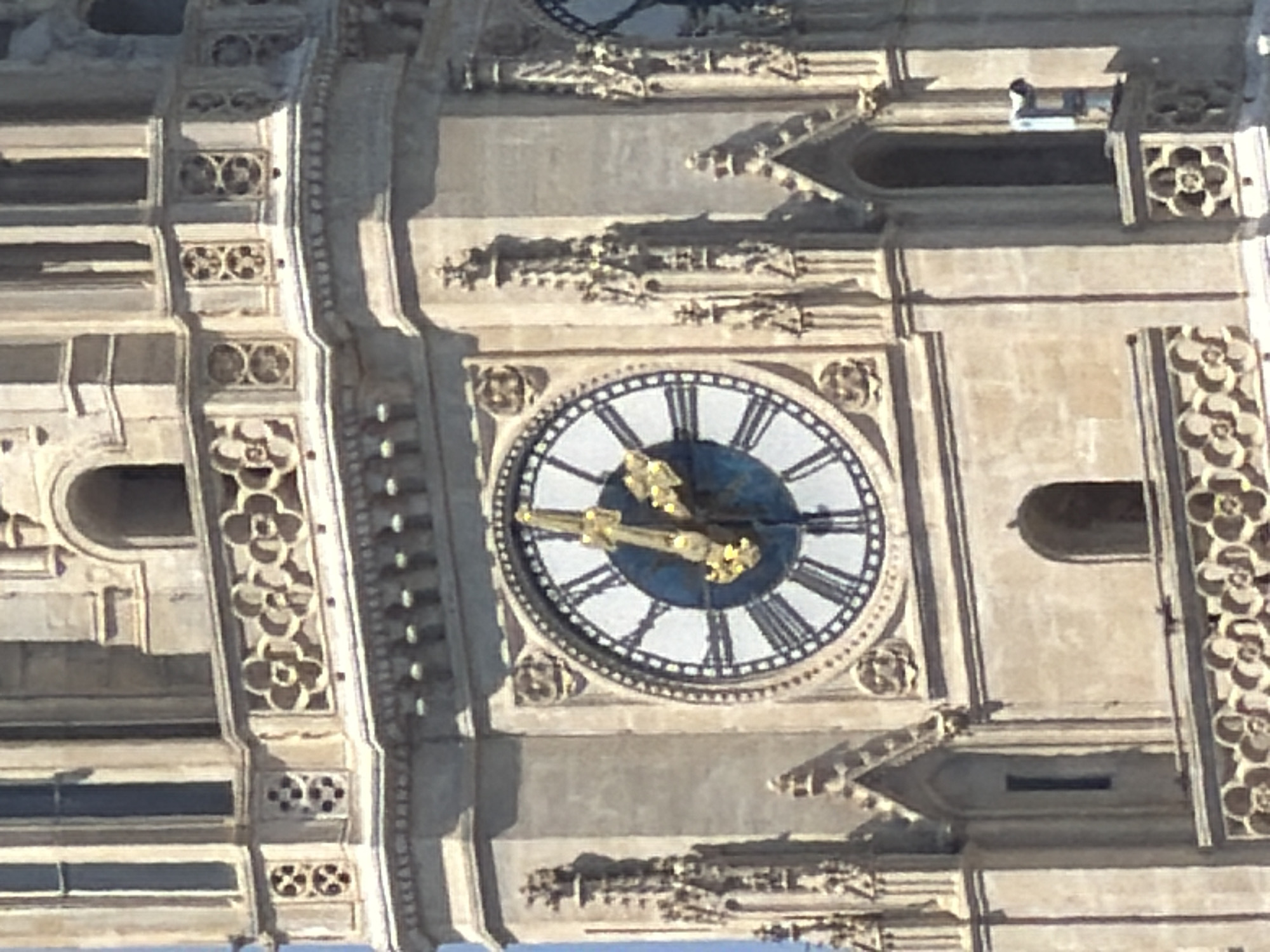 This is a close-up photograph of the clock tower of St. Stephen's Cathedral (Stephansdom) in Vienna, Austria. The image prominently features a large, ornate clock face with Roman numerals and ornate, golden hands. The hour hand is pointing slightly past the '9', and the minute hand is pointing towards the '12'. The clock face is set against a dark blue background. The surrounding architecture is of Gothic style, characterized by intricate stone carvings, pointed arches, and decorative tracery. The stone appears weathered and light brown or beige in color. Sunlight casts shadows on the building, highlighting the architectural details. The overall impression is one of historical grandeur and architectural artistry.