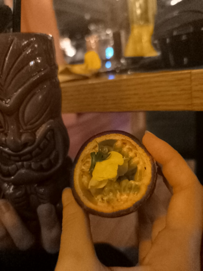 The image shows a dimly lit close-up of a person's hands holding a halved passion fruit, likely in a bar or restaurant setting in Palaio Faliro, Greece. The passion fruit, cut to expose its vibrant yellow-orange pulp and dark seeds, appears to be garnished with a small green leaf or herb and a light-colored, possibly edible, decorative piece. To the left, a dark brown tiki mug with a sculpted face, featuring pronounced eyes, nose, and a toothed mouth, stands upright. A dark straw or stirrer extends from the mug. The background is out of focus, revealing a wooden surface, possibly a bar counter, and blurred glints of light and yellow objects, which could be other drinks, bottles, or decor. The low lighting suggests an evening or nighttime setting. No visible text is present.