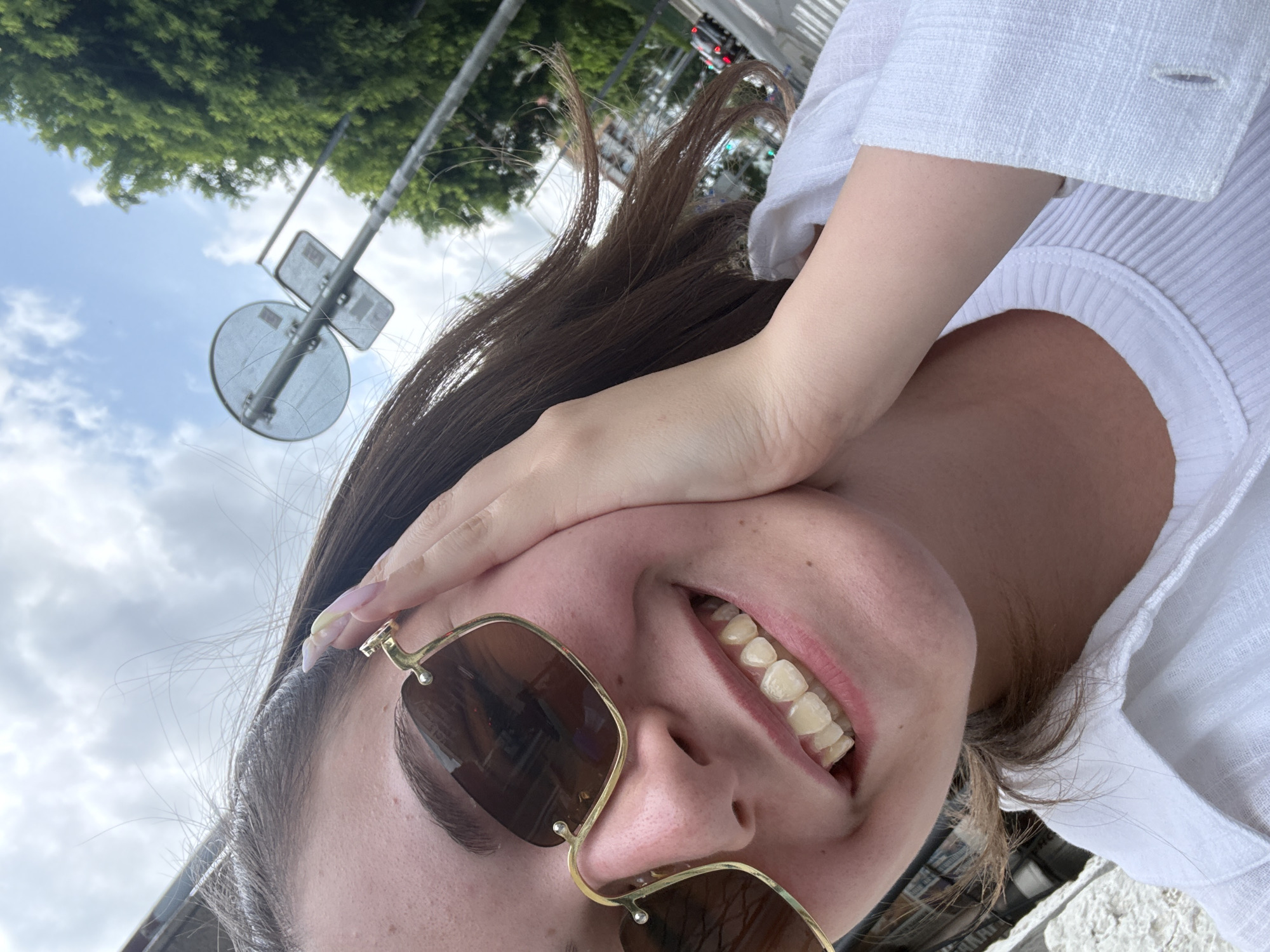 A young woman with dark brown hair and light skin is smiling directly at the camera, holding her right cheek with her left hand. She is wearing oversized gold-rimmed sunglasses with brown lenses and a white collared shirt over a white ribbed top. Her fingernails are painted with a pastel gradient. She has a cheerful expression and her teeth are visible. The background is slightly blurred, showing a cloudy sky, green trees, a street sign, and a street with some traffic, suggesting an outdoor urban setting. The image appears to be taken in Burgas, Bulgaria.