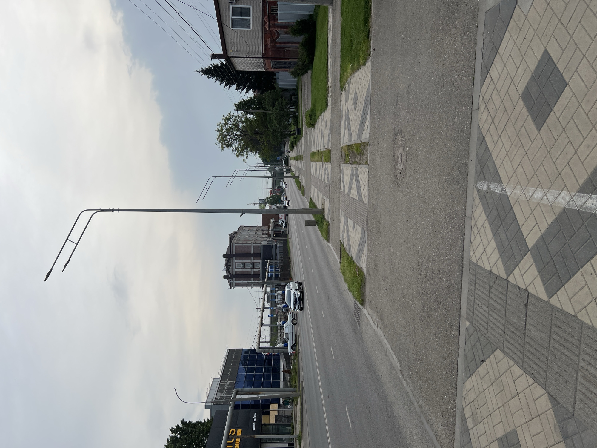 The image shows a street scene in Krasnodar, Russia. The camera is positioned along the side of the road. A wide sidewalk is on the right side, paved with alternating gray and light gray patterned paving stones. The road runs in the foreground to the far distance. A row of streetlights with curved arms line the road. Buildings of varying heights and styles are present on both sides of the street. Cars and white vans are driving on the road. The sky is overcast. A manhole cover is visible on the right side.