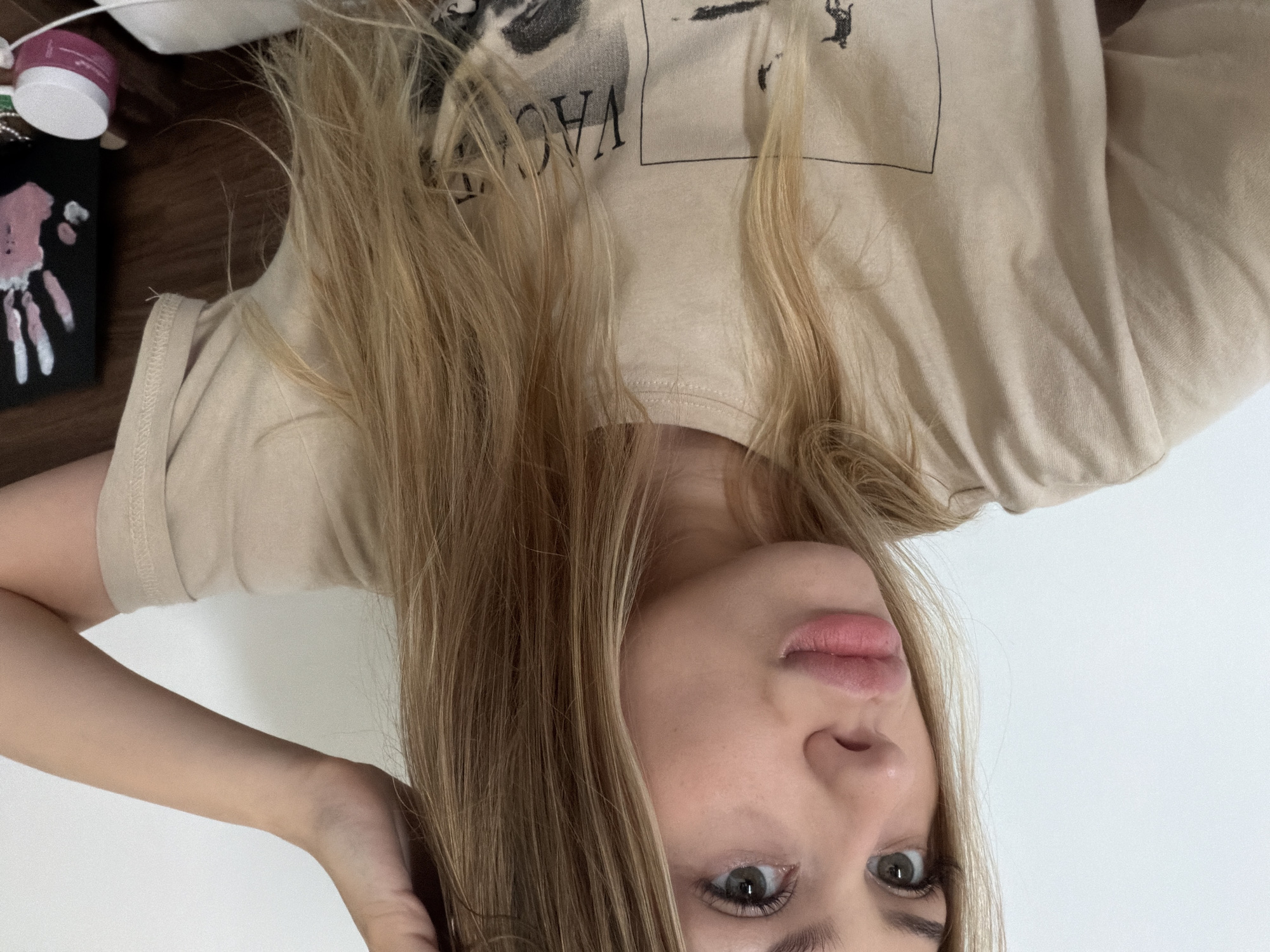 A young woman with blonde hair is pictured from the shoulders up, looking to her left with her right hand raised to touch her hair. She is wearing a tan t-shirt with a graphic print. The background is a plain white wall and a wooden surface. There are no identifiable landmarks or clues to suggest the location is Đà Nẵng, Vietnam.
