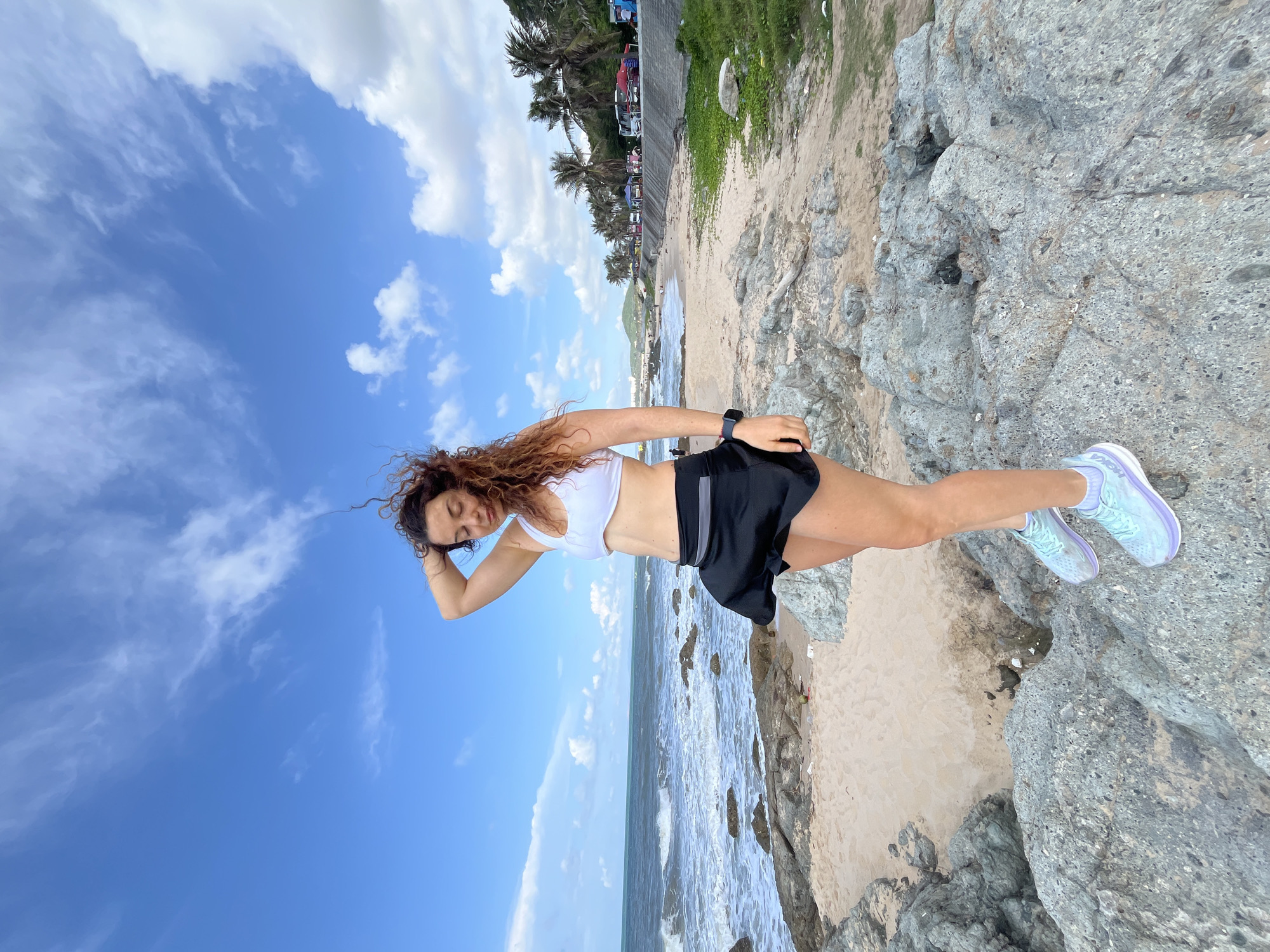A woman with curly brown hair stands on a rocky shore with the ocean to her left and a sandy beach to her right. She is wearing a white sports bra and a black skort, with light blue athletic shoes. Her right hand is raised, touching her hair, and her head is tilted down. The sky is blue with scattered white clouds. In the background, palm trees line the beach, and a road with parked vehicles and tents is visible. The location is Phu Thuy Ward, Vietnam.