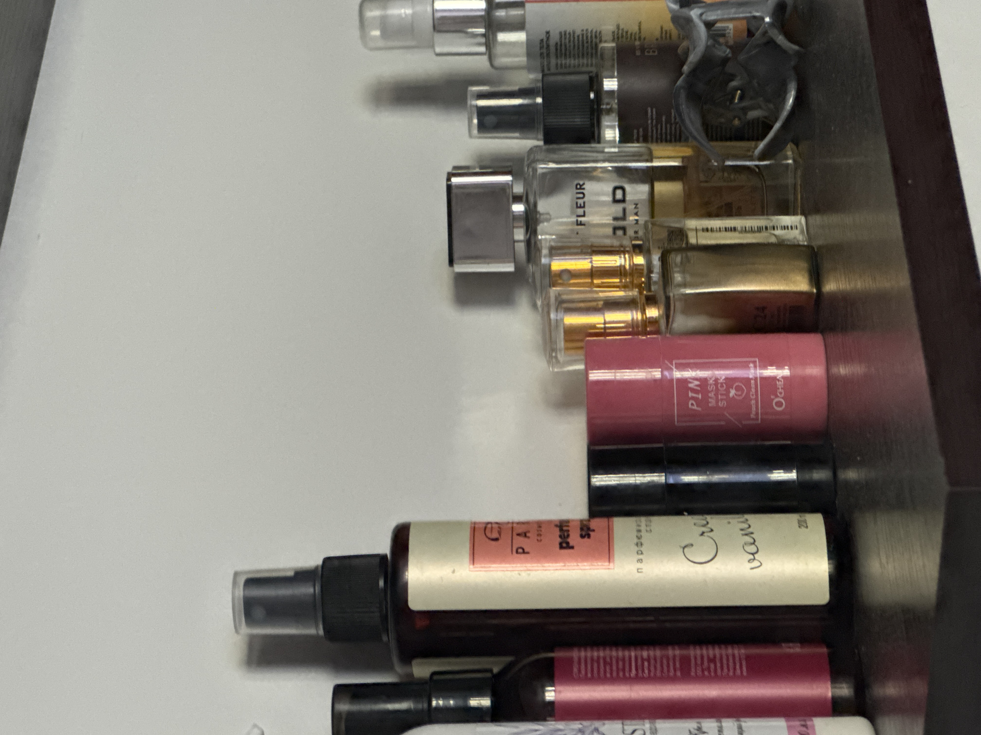 This media file is a close-up, eye-level shot of a collection of cosmetic and fragrance products, likely stored in a drawer or on a shelf. The items are arranged vertically.
In the foreground, there is a large spray bottle with a beige label that reads "PA COSMETICS" and "perf" and "spn". Next to it, partially visible, is another spray bottle with a label that includes the words "Cre" and "vanil" in a handwritten font. Below these are two more cylindrical tubes, one black and one with a pink label.
Above these are several smaller glass bottles, some with gold accents and clear spray tops, and others that appear to be smaller perfume bottles with decorative caps. One clear bottle in the upper middle has the text "FLEUR" and "OLD" visible. Further up, a black spray bottle is visible, and at the very top, a clear spray bottle with a silver cap. One item in the upper right quadrant appears to be a grey hair clip or accessory.
The background is a plain, light-colored surface, possibly a drawer liner, with a dark edge on the right side suggesting the interior of a cabinet or drawer. The lighting is neutral, indicating an indoor setting.
The visible text indicates a variety of cosmetic products, including perfumes and possibly skincare items, with brand names like "PA COSMETICS," and labels suggesting scents like vanilla. Some text is in Cyrillic, indicating a potential location in a region where this script is used, such as Belarus. There are no people or animals visible, and the focus is entirely on the arrangement of these personal care items. The overall impression is one of organized storage of beauty products.