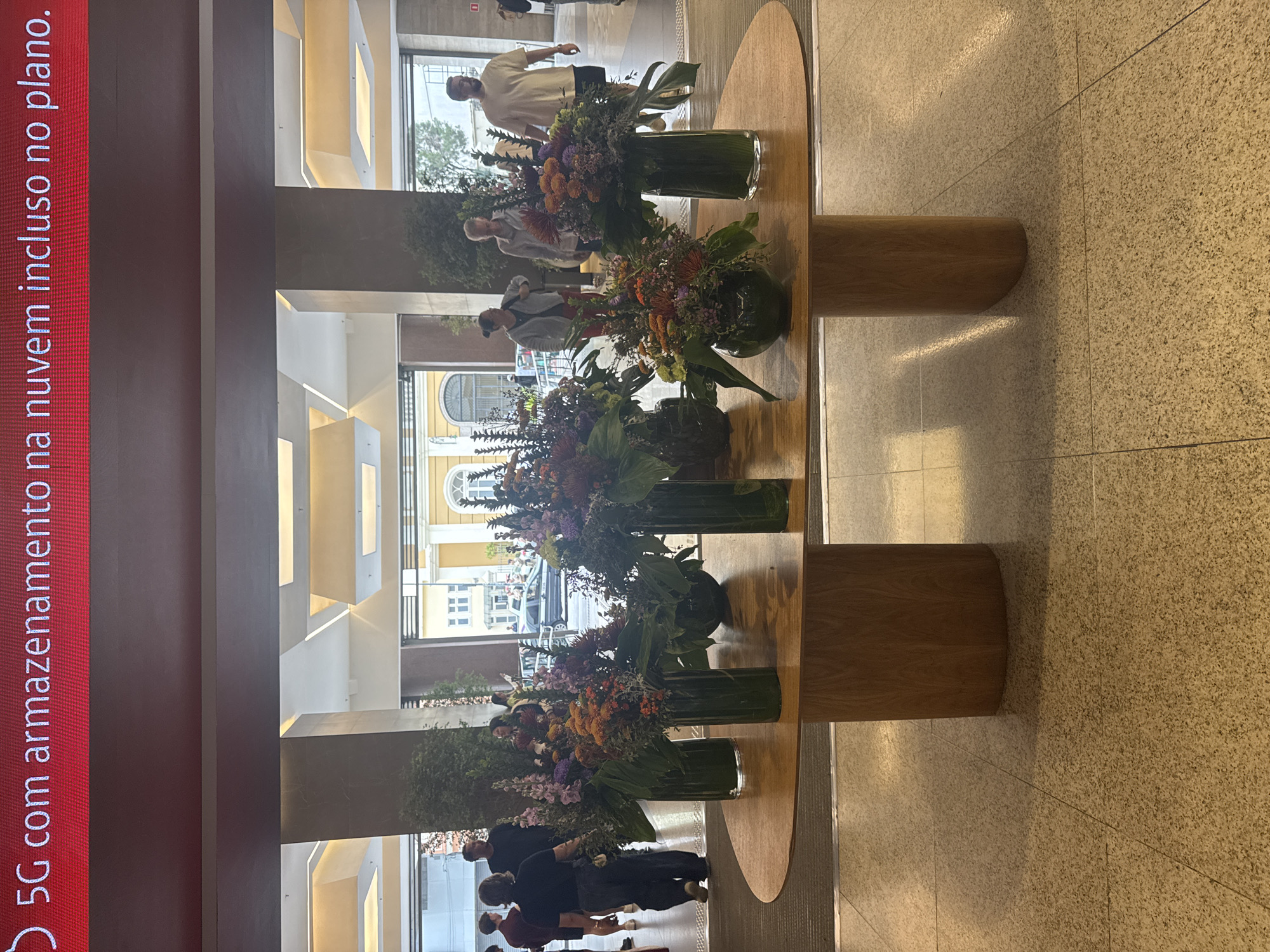 The image shows a wide shot of an indoor foyer area. At the top of the image, a red digital billboard displays white text that reads "5G com armazenamento na nuvem incluso no plano." The billboard spans the width of the image and is situated above a dark brown ceiling panel. Below this, a brightly lit atrium-like space is visible with recessed rectangular ceiling lights. In the foreground, a large, oval-shaped wooden table with rounded edges sits on the speckled beige tile floor. The table is supported by two thick, wooden, cylindrical legs.
Several large floral arrangements are placed on the table in clear glass vases. The bouquets are dense and feature a variety of colorful flowers and greenery, including orange, purple, and green elements. Some of the vases are tall and cylindrical, while one in the center is shorter and more rounded.
In the background, through a large opening, people are seen walking in what appears to be a street or public area. Some individuals are walking towards the camera, while others are walking away. The architecture visible in the background suggests a European-influenced building with yellow facades and arched windows. The setting appears to be a public space, possibly a commercial building, a hotel lobby, or a large retail establishment in Niterói, Brazil, given the Portuguese text on the billboard. The overall lighting is bright, highlighting the floral displays and the polished floor.