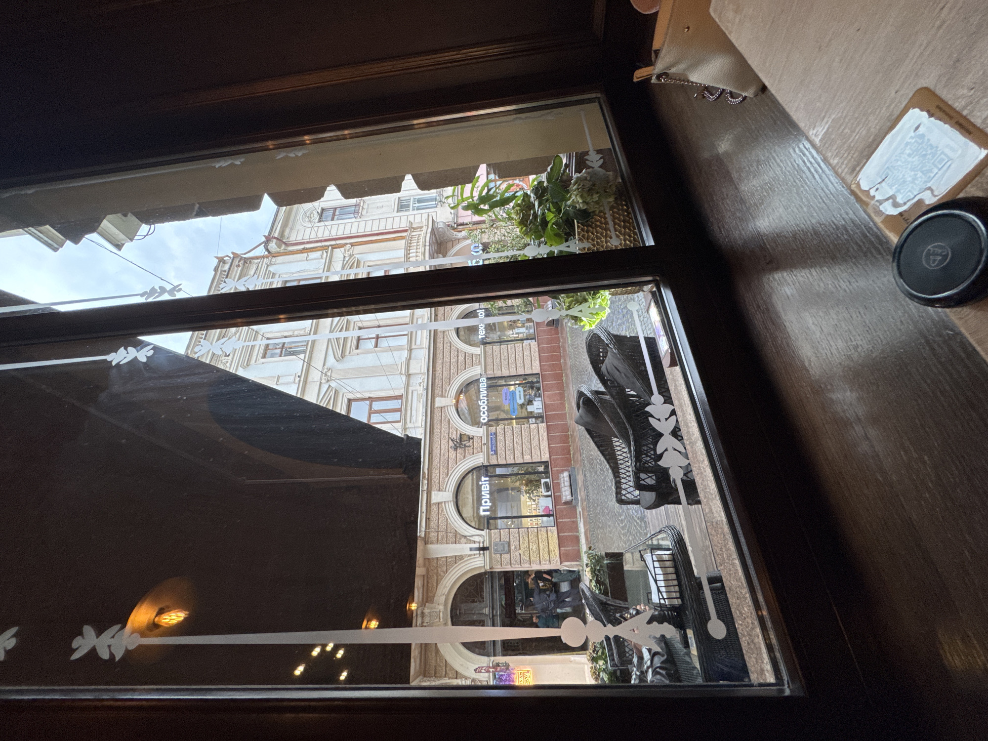 The image is a view through a window, likely from inside a cafe or restaurant, looking out onto a street scene in Chernivtsi, Ukraine. In the foreground, on the right side of the frame, a wooden table surface is visible with a small, round black object and a card with a QR code on it. To the left, through the window, an outdoor seating area with wicker chairs and tables is visible, set on a cobblestone pavement. Beyond the seating area, a row of shops lines the street. One shop has a sign that reads "Привіт" (Privit, meaning "Hello") in large letters, and another has a sign reading "особлива" (osoblyva, meaning "special"). The buildings across the street are multi-story and appear to be of historical architecture. A street lamp with an orange glow is visible reflecting in the window. The sky is overcast.