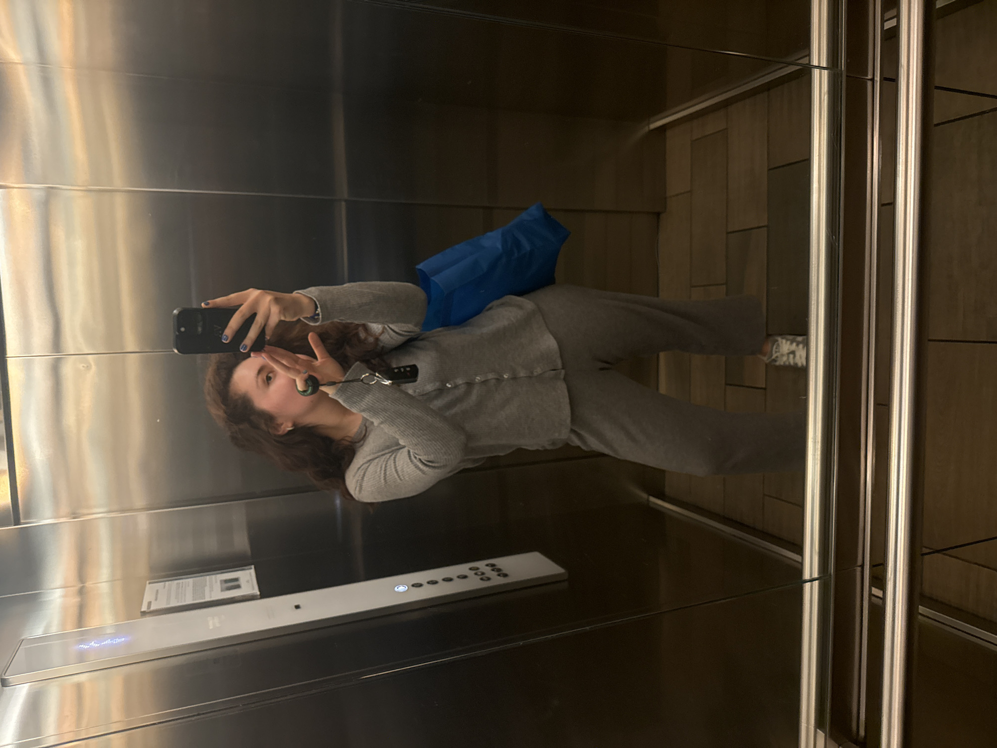 A young woman is standing in the mirrored interior of an elevator, holding a smartphone in her left hand to take a picture of herself. She is wearing a grey sweat suit and has a blue shopping bag on her lap. The elevator has stainless steel walls, a control panel with numbered buttons, and a sign. The floor appears to be tiled. The location is likely in Mexico City, Mexico.