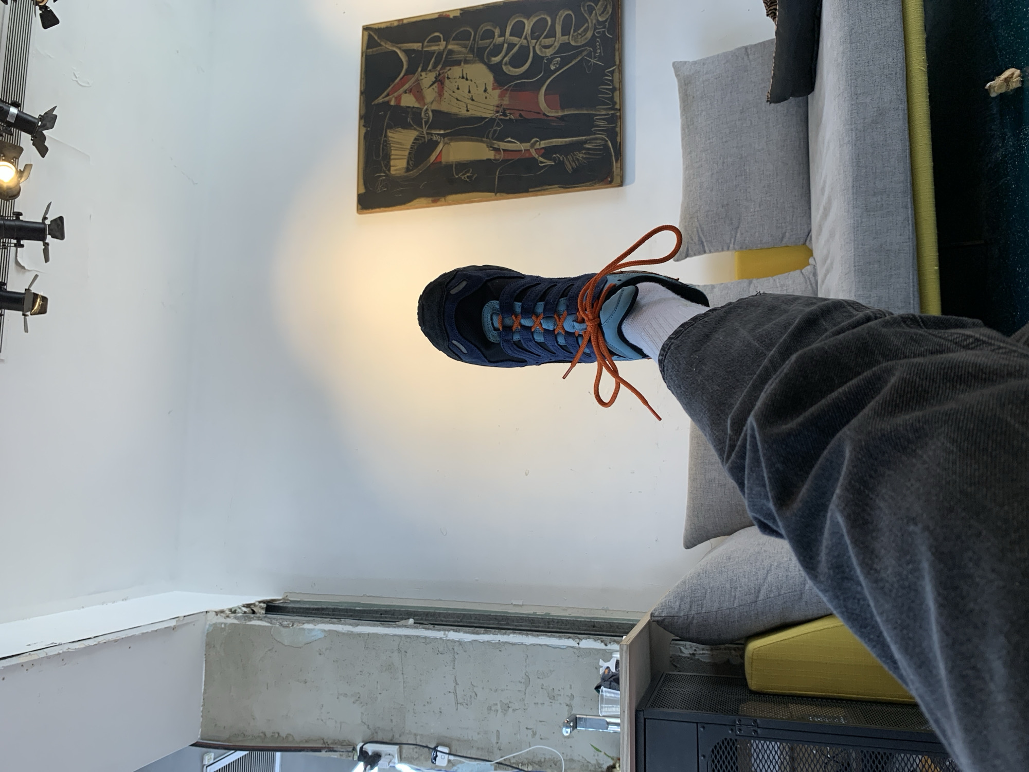 A person's leg, clad in grey denim jeans and a white sock, is angled towards the viewer, showcasing a navy blue and teal athletic shoe with orange laces. The leg extends from a grey couch. Above the couch and the shoe is a black, abstract painting hung on a white wall. A lighting fixture is visible on the left edge of the frame. In the lower portion of the image, the construction of the building is exposed, with bare concrete and electrical fixtures. The exact location in Tel-Aviv, Israel, cannot be determined from the information provided.