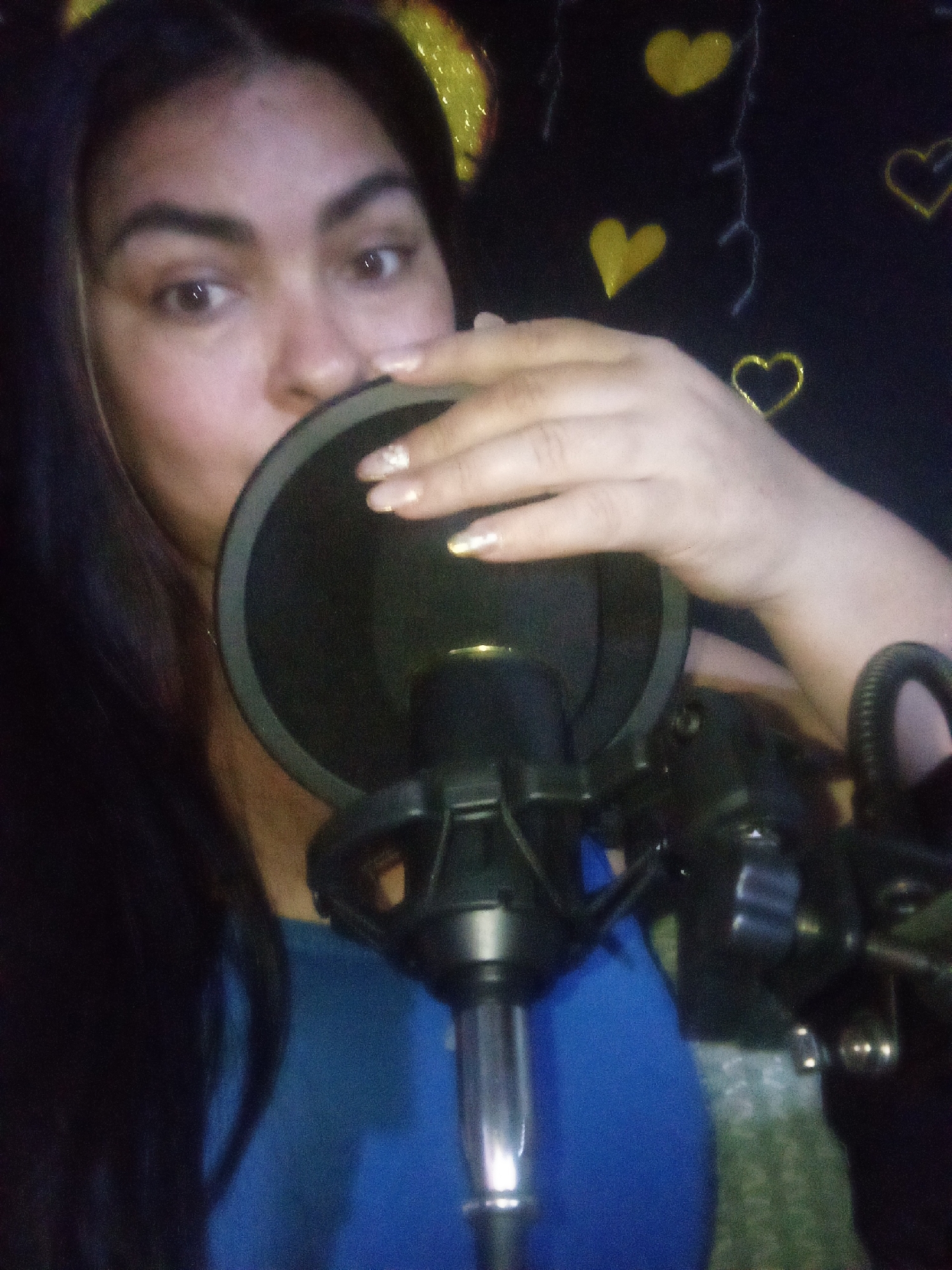 An indoor scene in Juan Griego, Venezuela, depicts a female subject positioned in front of a recording microphone setup. The subject has long, dark hair and wears a blue garment. Her right eye is open and directed towards the camera. Her right hand, featuring nails with glitter polish, is positioned near a black circular pop filter. Immediately behind the pop filter, a black condenser microphone is visible, mounted on a stand with a shock mount. A segment of a black headset is present on the right side of the frame. The background is a dark wall adorned with multiple yellow heart-shaped decorations, some solid and some outlined, interspersed with thin white linear patterns. The scene is illuminated by low to moderate lighting.