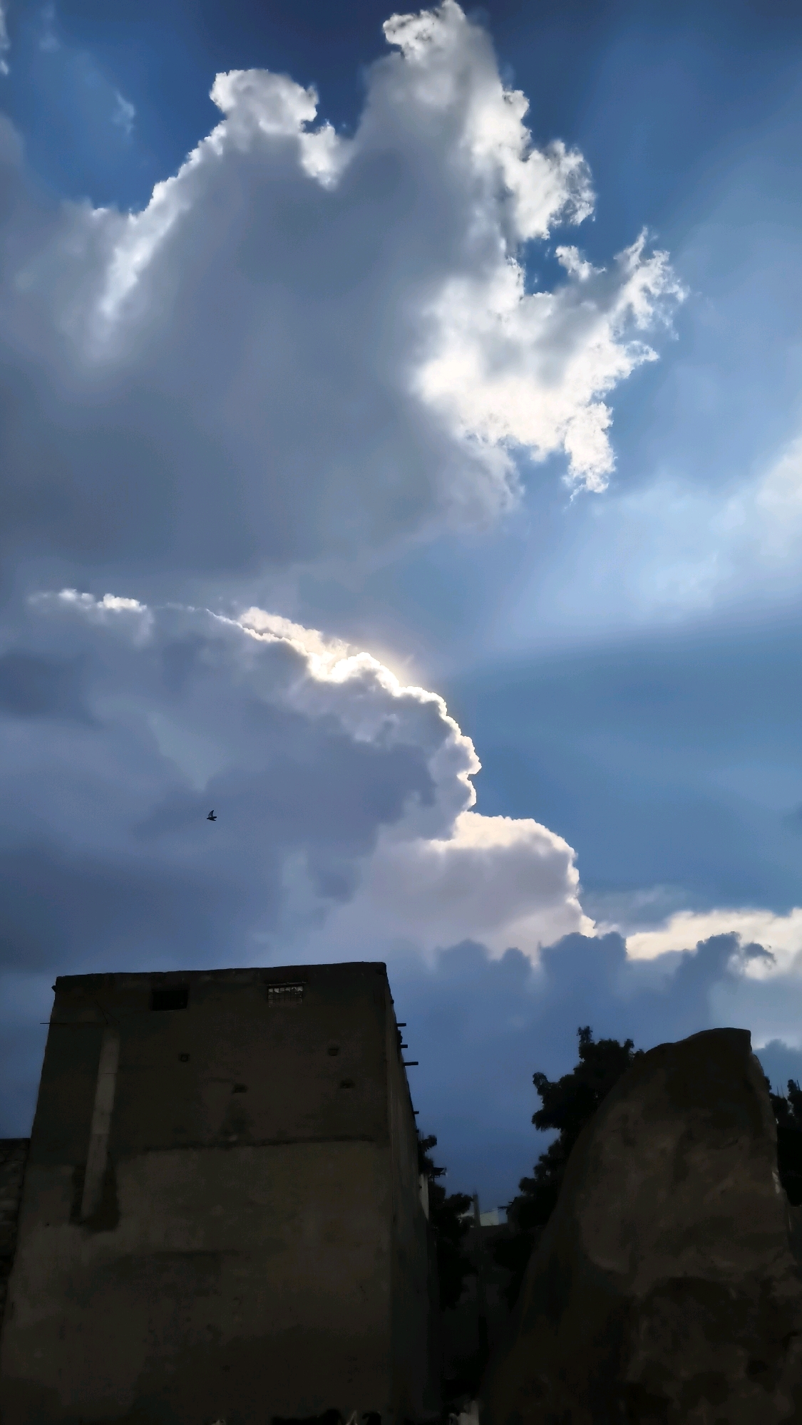 The image depicts a dramatic sky over what appears to be an urban or semi-urban environment. Large, voluminous clouds dominate the upper portion of the frame, with sunlight peeking through them, creating rays of light. The sky is a deep blue, transitioning to darker shades as the clouds thicken. In the lower part of the image, a silhouetted building with a rough, weathered facade stands prominently on the left. It has visible openings and some protruding wooden elements. To the right of the building, a large, irregular rock formation or structure is also silhouetted against the sky, with dark foliage of trees visible behind both structures. A small bird is captured in flight against the backdrop of the clouds. The overall lighting suggests either late afternoon or early evening, with the sun partially obscured by the clouds, creating a sense of atmospheric intensity. The image does not provide explicit visual cues to confirm the location as Hyderabad, India, but the architectural style of the building and the type of vegetation could be consistent with certain areas of the city.