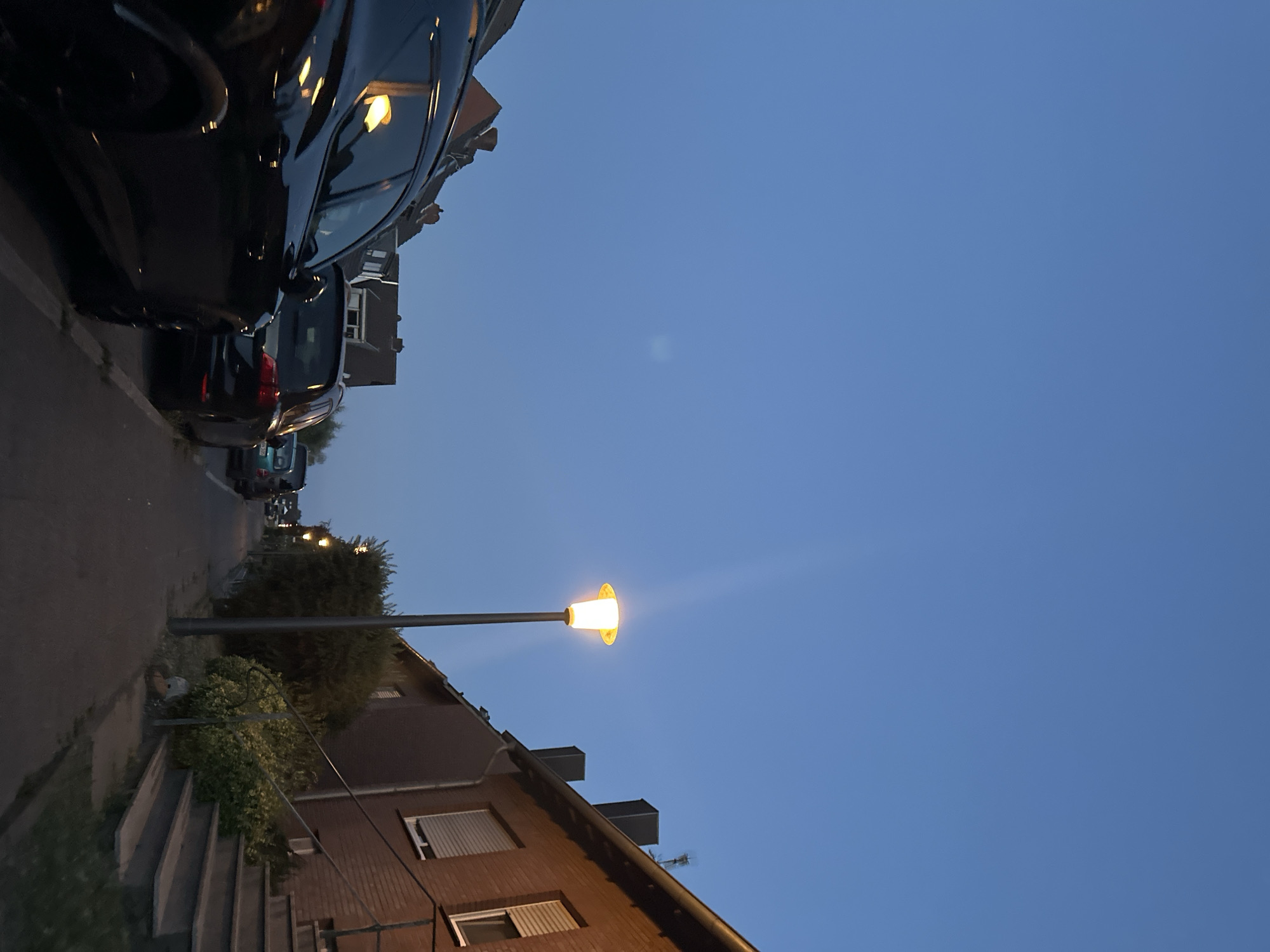 The image depicts a street scene at dusk in Krefeld, Germany. The sky is a clear, pale blue, indicating early evening. The foreground is dominated by the side of a dark-colored SUV parked on the left side of the street. Further down the street, several other cars are parked. To the right, a brick house with a tiled roof and closed shutters lines the sidewalk. A single lamppost with an illuminated yellow light stands on the sidewalk, casting a glow on the pavement. Dense green bushes and trees are visible between the house and the street. The overall impression is a quiet residential street transitioning into nighttime.