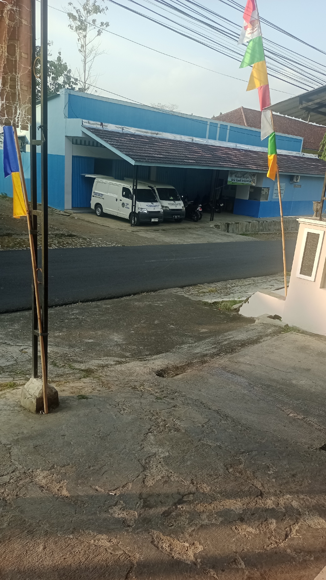 In Karyamandala, Indonesia, a two-story building with a bright blue facade and a brown-tiled overhang features two white vans parked beneath the shelter. A motorcycle is also visible under the overhang. Two flags, one with blue and yellow sections and another with red, white, green, and yellow sections, are displayed on poles in front of the building. The scene is set against a backdrop of trees and power lines, with a paved road and concrete ground in the foreground. The sun is casting shadows, suggesting it is either early morning or late afternoon.