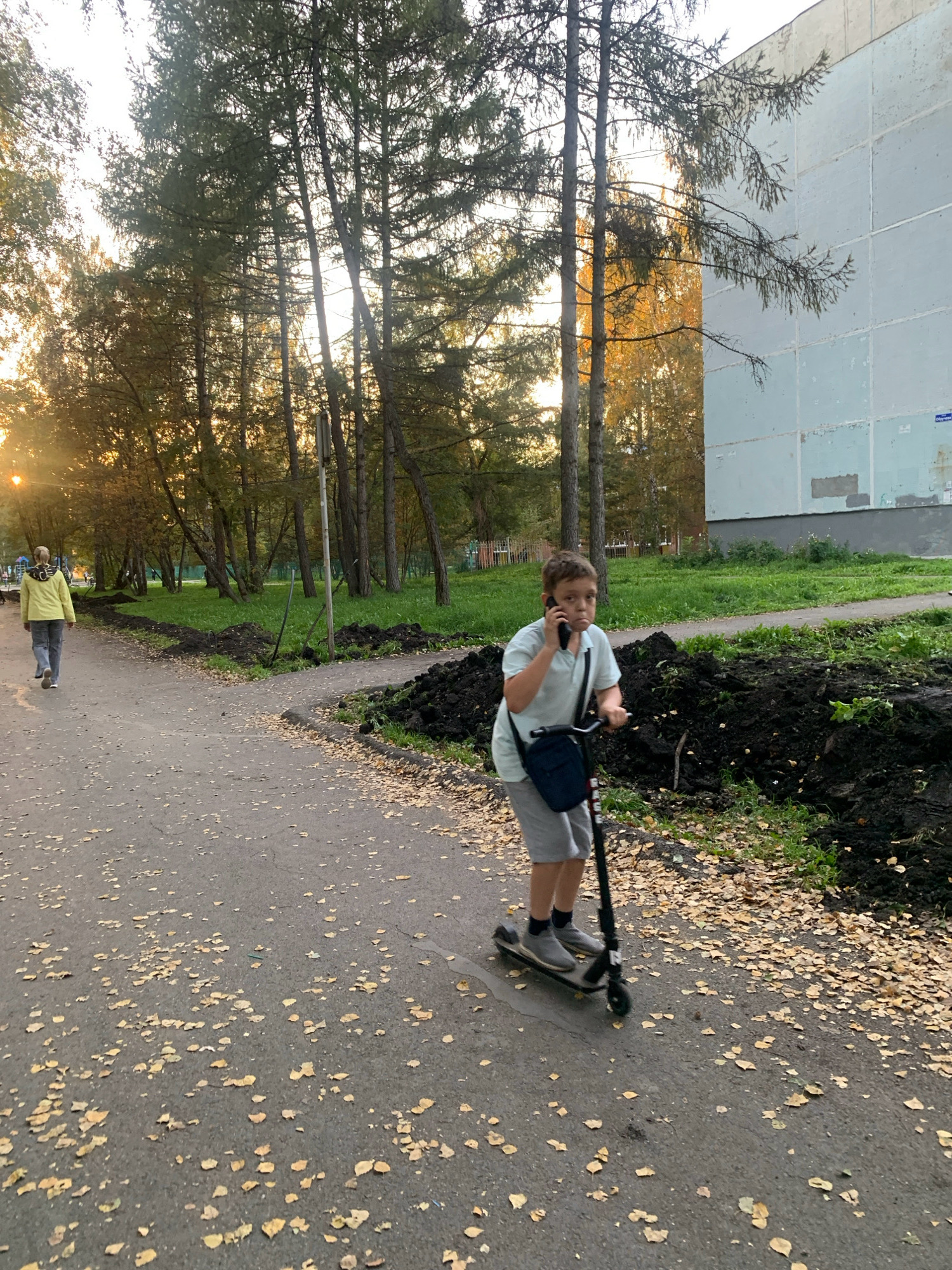 A young boy is riding a scooter and talking on his phone in the city of Prokopyevsk, Russia. He is wearing a light blue polo shirt and gray shorts. A dark blue bag is slung across his chest. He is riding on a paved path that is covered with fallen yellow leaves. To his right, there are piles of dark soil and green grass. Behind him, a woman in a yellow jacket is walking away from the camera. In the background, there are tall trees and a light blue building. The sun is setting, casting a warm glow on the scene.