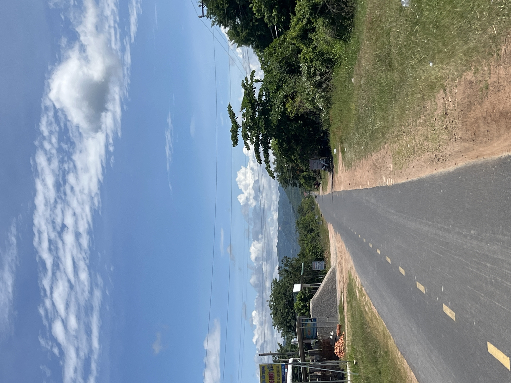 The image depicts a rural road in Ham Thuan Bac, Vietnam, on a partly cloudy day. The road is paved with asphalt and has yellow dashed lines marking the lanes. On the left side of the road, there is a small construction site with stacks of bricks, piles of gravel, and some makeshift shelters. A sign with Vietnamese writing is also visible. On the right side of the road, the terrain is covered with grass and scattered debris. Lush green trees and vegetation line both sides of the road, with rolling hills and mountains visible in the distance. Power lines are strung across the sky. A white motorcycle is parked on the side of the road near the construction area. The sky is a vibrant blue with scattered white cumulus clouds.