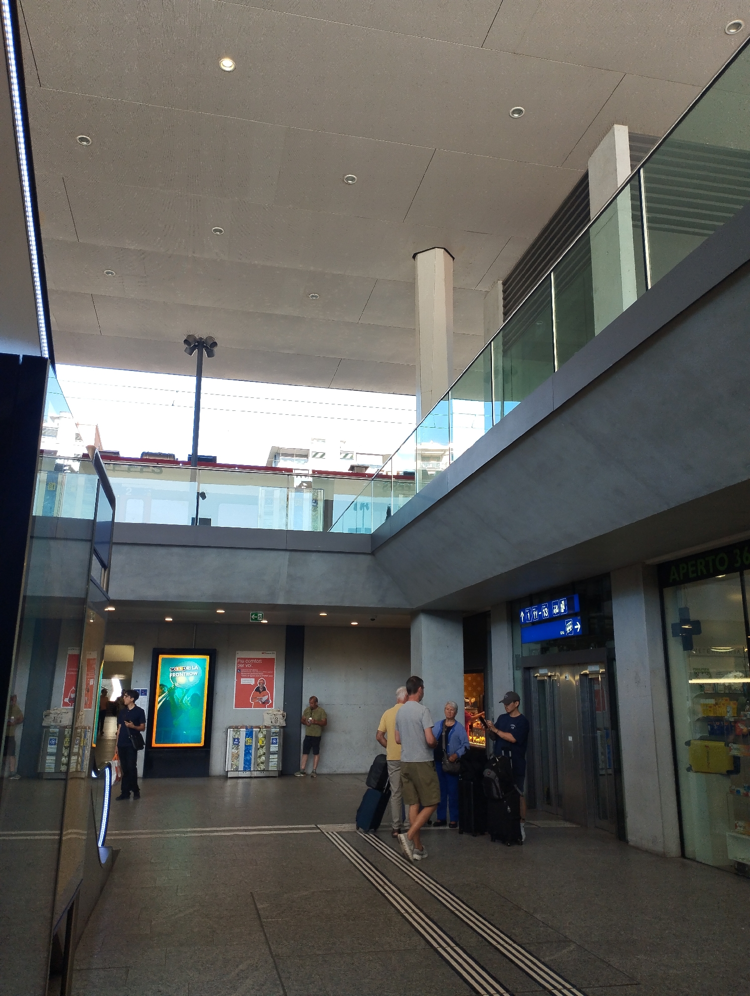 This image captures a scene at the Lugano train station in Lugano, Switzerland. In the foreground, a reflective glass wall dominates the left side of the frame, mirroring the environment. Several individuals are present: a person in a dark uniform stands near a large illuminated advertisement, another person in a blue jacket and grey pants walks away from the camera, and a group of three people, two men and a woman, are gathered with luggage near an elevator. The group appears to be waiting or interacting.
The environment suggests an indoor public space, likely a station concourse, with modern architecture. A suspended ceiling with integrated lighting is visible overhead, along with concrete pillars and a glass balustrade of an upper level. There are also signs, including an illuminated advertisement and directional signs with symbols and text indicating platform numbers and directions. The floor is tiled with a pattern of grey squares and white lines. In the background, storefronts and buildings are visible, hinting at the commercial activity within or adjacent to the station. The overall lighting is bright, suggesting daytime.