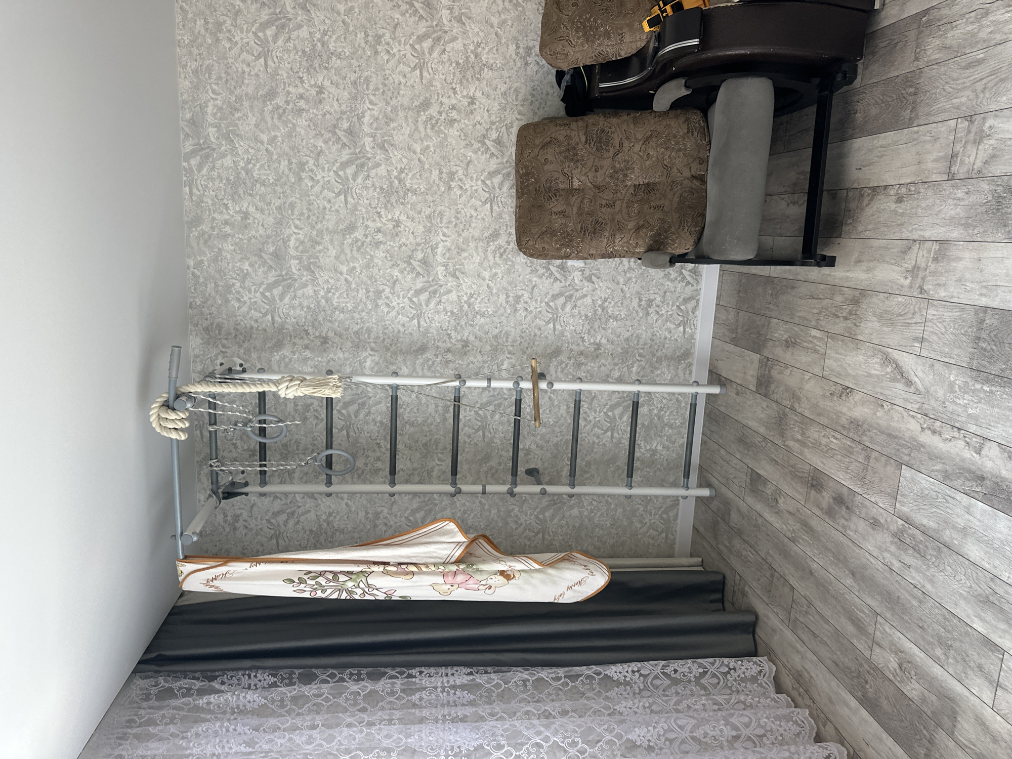 The image depicts an indoor scene, likely a living room or playroom, with a focus on a wall-mounted gymnastics apparatus. The apparatus is white with dark gray rungs and features various attachments, including rings, a rope with a thick knot, and a small wooden plank. A light-colored blanket with a cartoon bear print is draped over one side of the apparatus.
To the left of the gymnastics equipment, there are two sets of curtains: a sheer white lace curtain layered over a dark gray opaque curtain. The wall behind the apparatus is covered in a textured, light gray wallpaper with a subtle botanical pattern.
In the foreground and to the right, a dark brown recliner-style chair is partially visible. The chair has patterned cushions and appears to be in a reclined position. The flooring is a light gray laminate with a wood plank design. The overall impression is a well-lit, domestic space. The specific location in Mariinsky, Russia cannot be determined from the visual information provided in the image.