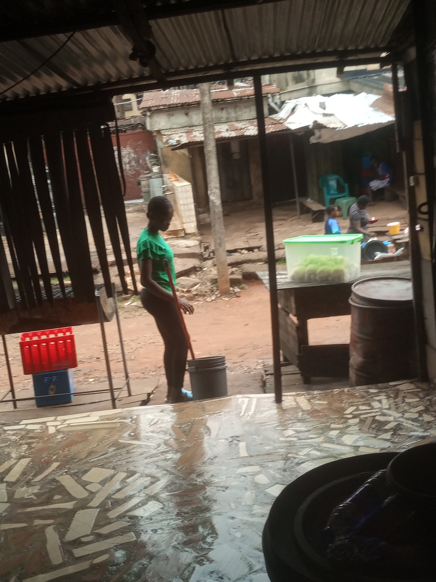 The image depicts a scene inside an open-air market or food stall, likely in a developing urban area, given the corrugated metal roof and rough construction. A young Black woman, wearing a bright green t-shirt and dark leggings, is the central figure. She stands with her back to the viewer, facing away from the camera, holding a red-handled broom or stick. She appears to be in the process of sweeping or cleaning. To her left, a red plastic crate filled with bottles is stacked on top of a blue crate. In the mid-ground, a wooden table holds a clear plastic container filled with pale green fruits, possibly avocados or guavas. To the right of the table is a large, dark metal barrel. The ground is wet and reflective, suggesting recent rain or cleaning. In the background, other individuals are visible, including a person in a blue shirt and another in a striped shirt, engaged in activity, though their actions are unclear. Buildings with corrugated metal roofs are visible in the distance, indicative of a densely populated area. The overall impression is one of a functional, albeit rustic, marketplace.