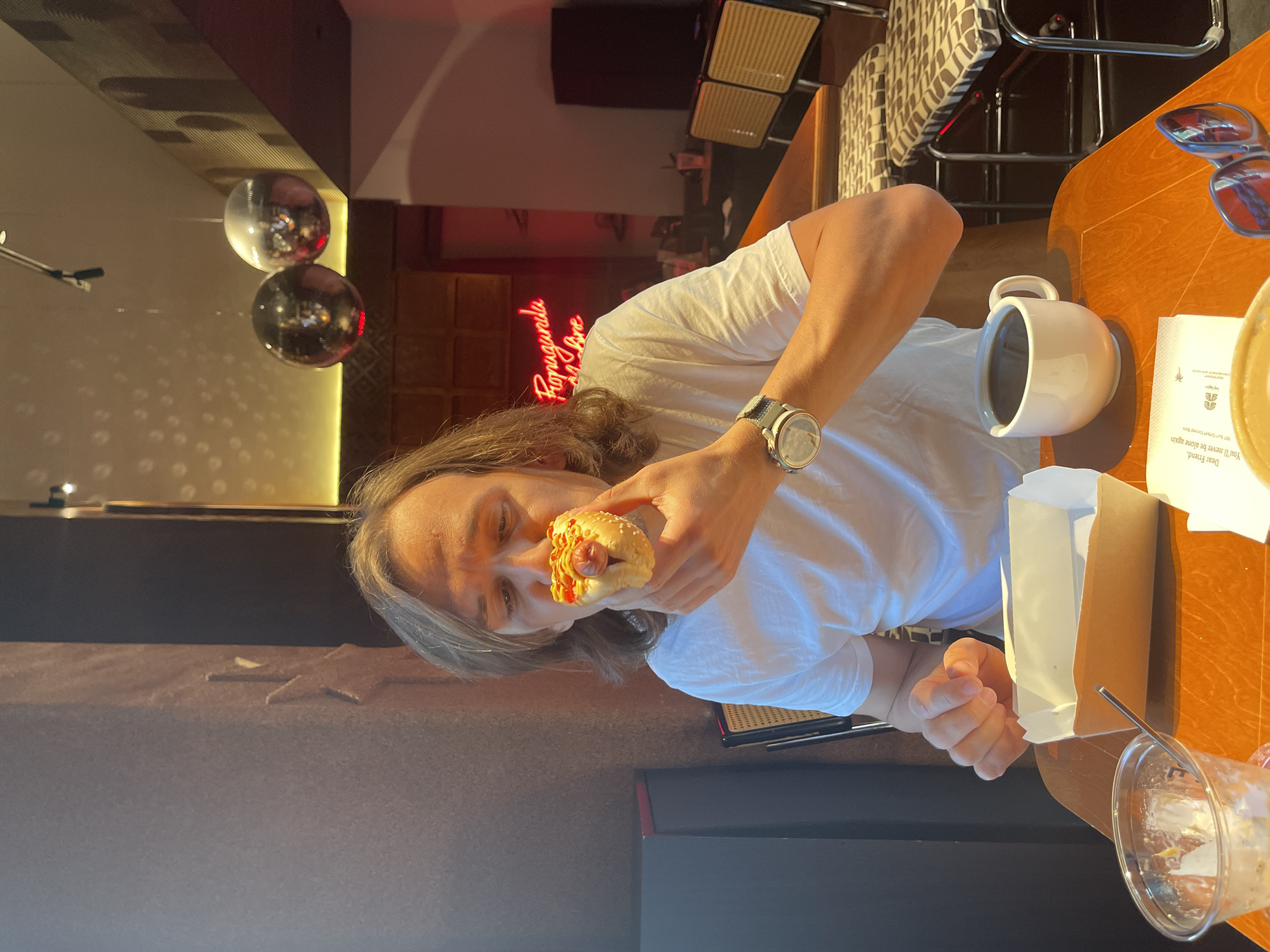 A man with shoulder-length hair and a watch on his left wrist is eating a hotdog in what appears to be a restaurant. He is wearing a white t-shirt. On the table is a cup of coffee, a white paper bag, an empty cup with a straw, and a pair of sunglasses. Above the man is a neon sign that says "Propaganda Izhe." This would suggest that the location is in Izhevsk, Russia. There are also two mirrored disco balls near the ceiling. Behind the man, there are chairs and other tables.