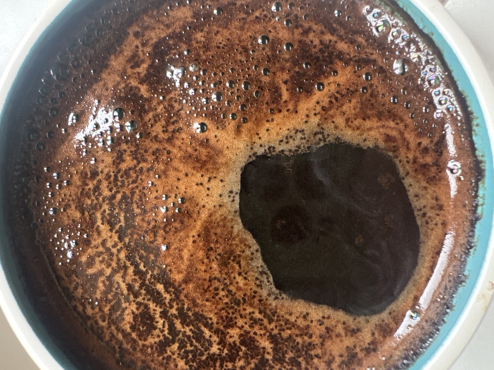 A close-up, top-down view captures a freshly prepared cup of coffee, likely in Moscow, Russia, during daytime. The primary subject is the rich, dark brown coffee, covered by a significant layer of reddish-brown crema. This crema displays an intricate, mottled texture with darker specks, possibly fine coffee grounds, distributed across its surface. Numerous small bubbles are visible within the crema; many of these bubbles, especially towards the edges of the cup, reflect light with iridescent, rainbow-like colors.
A distinct, irregularly shaped, darker area is present in the center-right of the cup, where the crema appears to have thinned or broken, revealing the deeper, darker liquid coffee beneath. The rim of the ceramic cup is partially visible, featuring a band of light blue or turquoise along its inner edge, complemented by a wider white or off-white outer section. The lighting is bright and appears natural, enhancing the reflections and textural details on the coffee's surface. No visible text is present in the image.