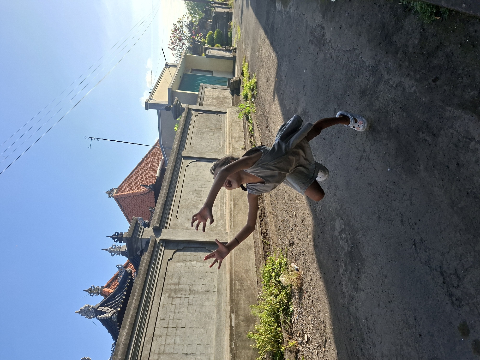In an outdoor, urban setting with a clear blue sky, a young girl in a grey dress and white shoes is captured mid-air, appearing to jump or fall. Her arms are outstretched and her mouth is open in an expression of excitement or surprise. She is positioned in the center of a paved road, with a grey concrete wall and tiled rooftops lining the left side of the frame. Green foliage sprouts along the base of the wall. On the right side of the road, a building with a light-colored facade and a teal door is visible, along with a tree with pink blossoms. Electrical wires stretch across the sky. The lighting suggests it is daytime, with shadows cast by the girl and the structures.