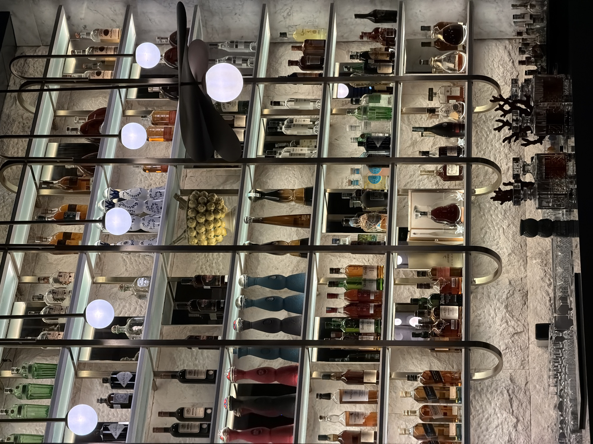 The original media file is rotated 90 degrees clockwise, depicting an upscale, brightly lit bar or liquor display as it would appear upright. The scene is set in a modern, sophisticated establishment, likely a bar or high-end lounge, possibly in Budva, Montenegro.
The primary subjects are numerous bottles of alcoholic beverages, neatly arranged on multiple tiers of glass shelves. These shelves are framed by sleek, metallic U-shaped structures and illuminated from within, casting a warm glow on the spirits. Several spherical white lights are integrated into the display at various levels, serving as both lighting and decorative elements. A prominent, dark, abstract, fan-like sculpture or decorative piece vertically spans several shelves on the left side of the display.
The bottles showcase a wide variety of spirits, including whiskey, gin, vodka, and more, identifiable by their distinct shapes, colors, and labels. Visible brands from the OCR and image include HENDRICK'S Gin, Glenfiddich, DALMORE, Ardbeg, Hennessey, Tito's Handmade Vodka, HOYO Club, and other labels like Jame, GLAY, RA, AME, and Hande.
The background wall behind the shelves features a light-colored, rough stone texture, adding an element of natural elegance to the modern design. On the far right, beyond the main shelving unit, there are additional decorative bottles, possibly decanters, and what appears to be a section of a bar counter with small glasses, further emphasizing the luxurious setting. There is no human activity visible in the image, focusing attention entirely on the meticulously curated display of beverages and the refined interior design.