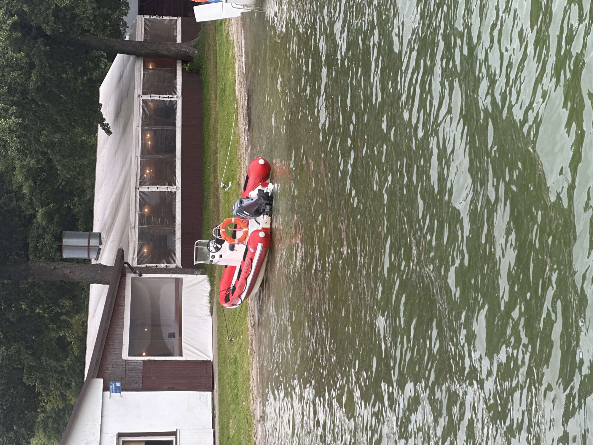 A red and white inflatable boat with a black outboard motor is tied to the shore of a body of water. The boat is positioned on the water's edge, with its bow facing left. A life preserver ring is visible on the boat's stern. In the background, there is a building with a white facade and a brown wooden door. Adjacent to this building is a covered outdoor seating area with clear plastic siding and lights inside. Trees and foliage form a dense green backdrop behind the structures. The scene is set on the shore of Lake Kłodno in Chmielno, Poland.