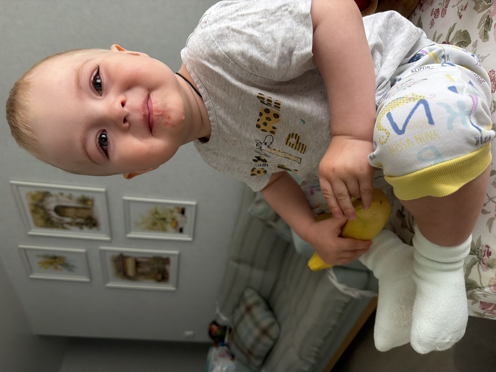 A young child is seated on a floral-patterned surface, possibly a table or couch, indoors. The child is fair-skinned, with blonde hair, and is wearing a light grey t-shirt with animal print designs, white shorts with yellow trim, and white socks. The child is holding a yellow object, possibly a fruit or toy, in their hands. Behind the child, there is a light grey wall with four framed pictures hanging on it, and a grey sofa with patterned cushions. Based on the visual elements, there is no indication that the scene is in Votkinsk, Russia, and that information is likely inaccurate.