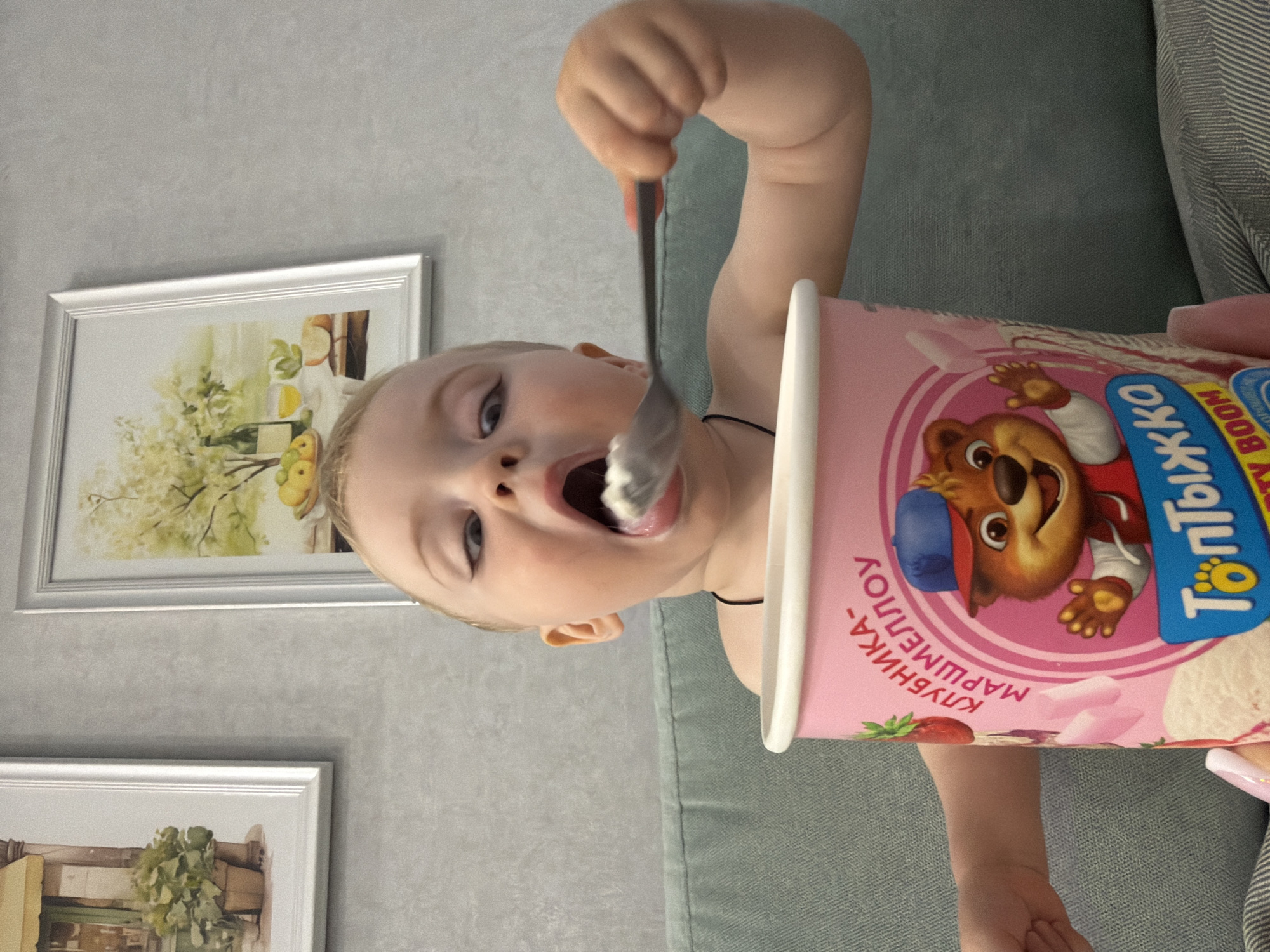 A young child is eating ice cream from a pink container with a cartoon bear on it, holding a spoon to their mouth. The text on the container is in Russian. The child is seated on a grey couch. Behind the child are two framed pictures on a light grey wall. The location is likely in Votkinsk, Russia, based on the text on the ice cream container.