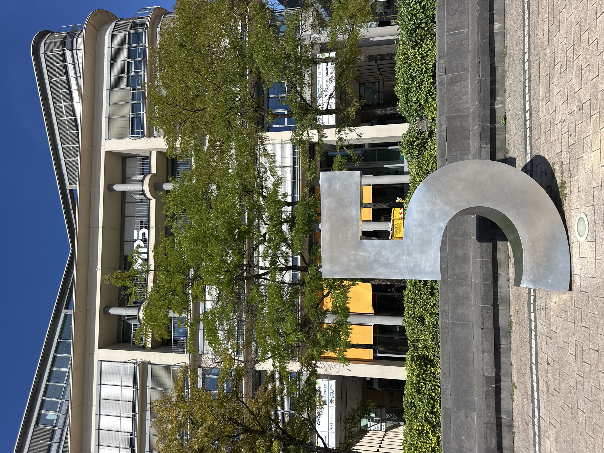 An outdoor, eye-level, full shot captures a large, metallic, freestanding number "5" sculpture in the foreground, made of brushed metal and casting a subtle shadow on the paved ground. Behind the sculpture, a contemporary building with a curved facade features multiple stories with windows covered by blinds and balconies with railings. Green trees with lush foliage partially obscure the building, their branches reaching towards the structure. Yellow awnings are visible above street-level entrances. A low, dark stone wall with a dark gray stone bench runs horizontally across the middle ground, separating the paved area from a dense hedge of green shrubs. The sky above is clear and blue, indicating a sunny day. The scene is set in front of the Cologne Tower (KölnTurm) in Cologne, Germany, identifiable by its distinctive architecture and the stylized "MPS" logo visible on the building's facade. No people are clearly visible.