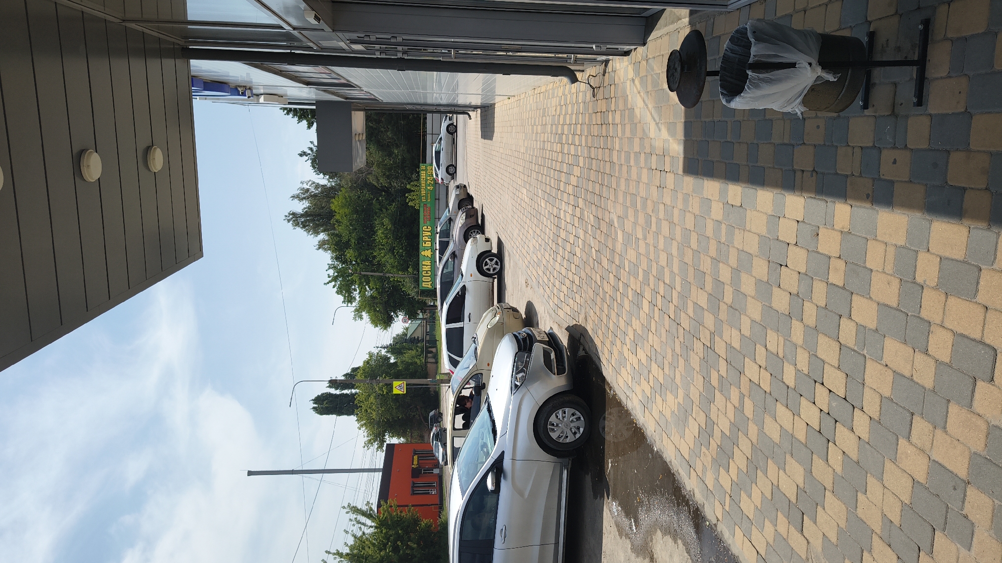 The image shows a parking lot and the exterior of a building in Mikhaylovka, Russia. In the foreground, a silver car is parked on a paved area. Behind it, several other cars are parked in a row, including a beige sedan and a white hatchback. The building's facade is visible on the right, with large windows and a grey, corrugated metal overhang. A trash can with a plastic liner stands near the building. In the background, trees and a blue sky with white clouds are visible. A sign with Russian text, including "ДОСКА БРУС" (plank and timber), is also present. The overall scene depicts a typical commercial area on a sunny day.