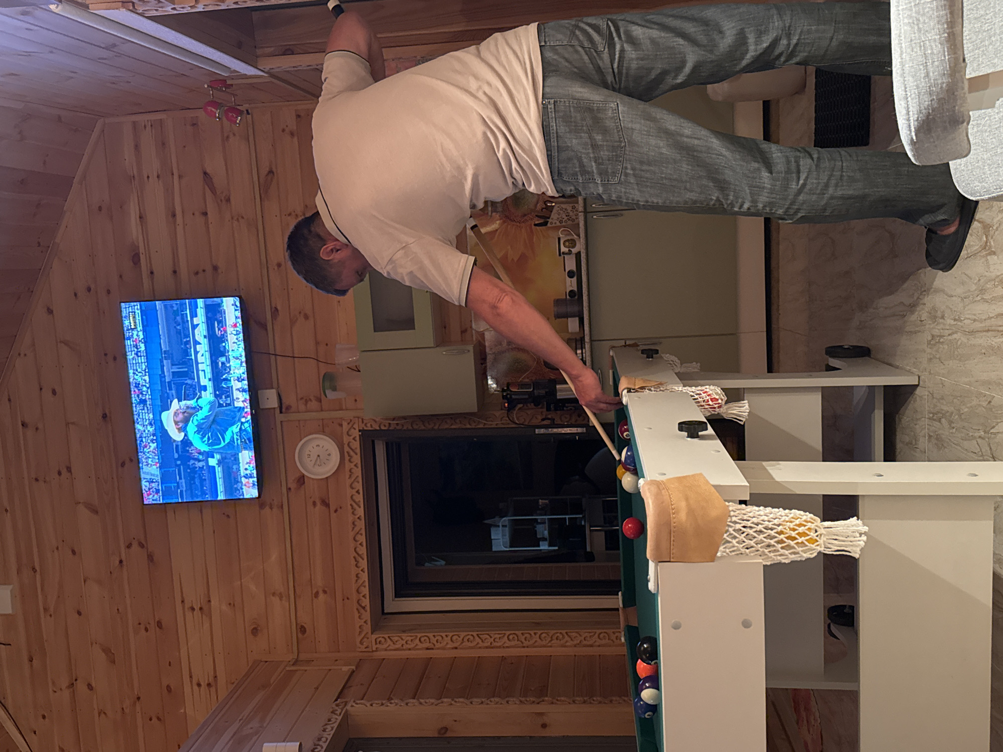 The image depicts an indoor scene, likely a recreational room or a cabin, with wooden paneling on the walls and ceiling. In the foreground, a man wearing a light-colored polo shirt and blue jeans is playing billiards. He is holding a pool cue and is in the process of taking a shot on a green felt pool table. The table is equipped with several colored billiard balls.
The room features a television mounted on the wall, displaying an image of a man in a cowboy hat. A clock is visible on the wall above a doorway. In the background, kitchen cabinets and a countertop are present, suggesting a combined living and kitchen area. The overall environment appears to be rustic and cozy, with wooden accents throughout. The specific location in the city of Zelenaya Roshcha, Russia, is not visually indicated in the image.