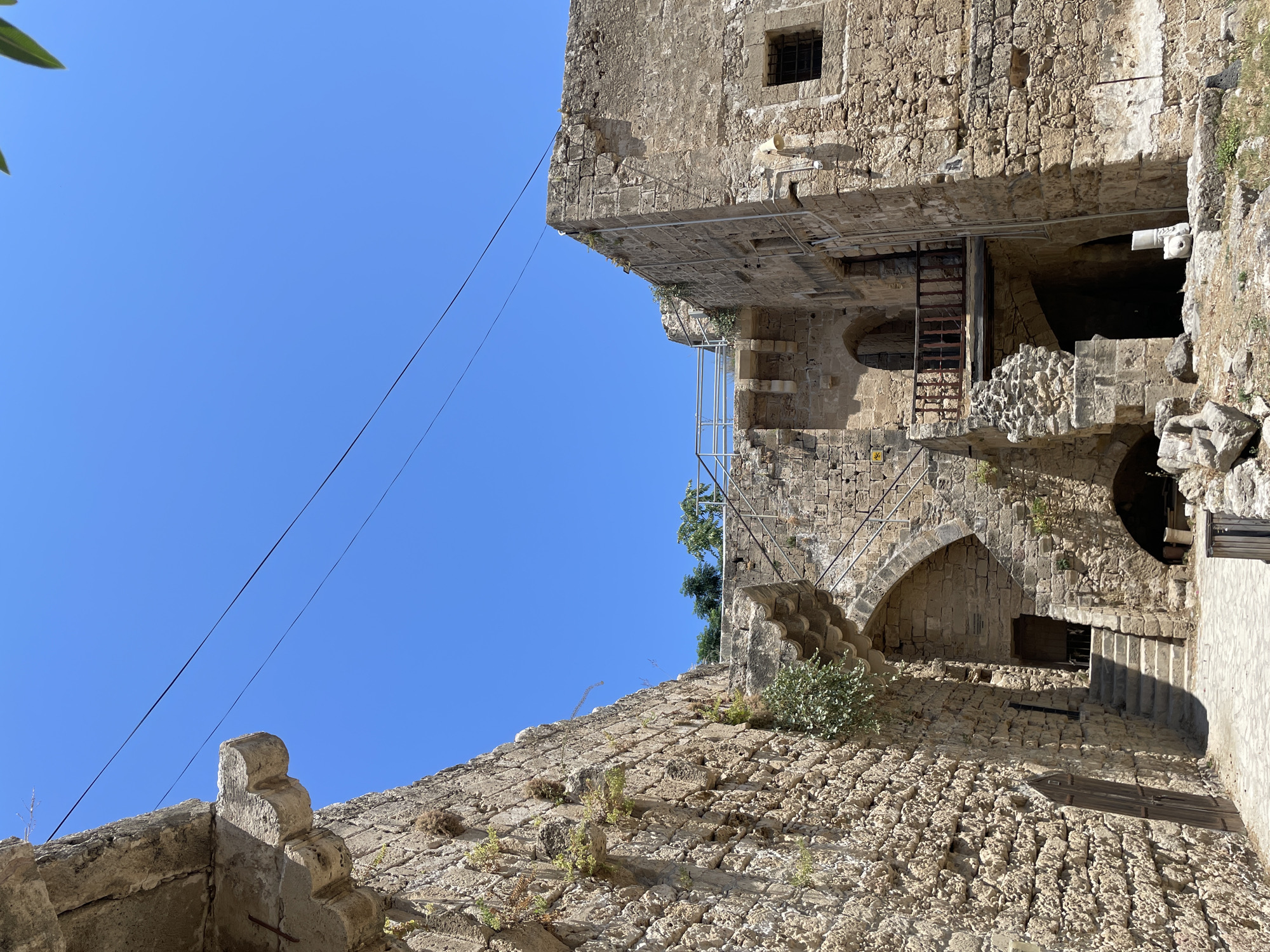 The image depicts the interior courtyard of Kyrenia Castle, a medieval landmark in Kyrenia, Northern Cyprus. The courtyard is surrounded by ancient stone walls, featuring arches, staircases, and balconies constructed from rough-hewn stone blocks. Vegetation, such as moss and small bushes, grows in the crevices of the stonework, indicating age and exposure to the elements. The sky above is a clear, vibrant blue, suggesting a sunny day. The ground is paved with cobblestones in the foreground, transitioning to dirt and debris in the background. A wooden door is visible on the left wall, and a set of stone stairs leads up to an arched entryway. A wooden balcony juts out from the right wall, above a dark, arched opening. The scene is devoid of any people, focusing solely on the architecture and the atmospheric quality of the historical site.