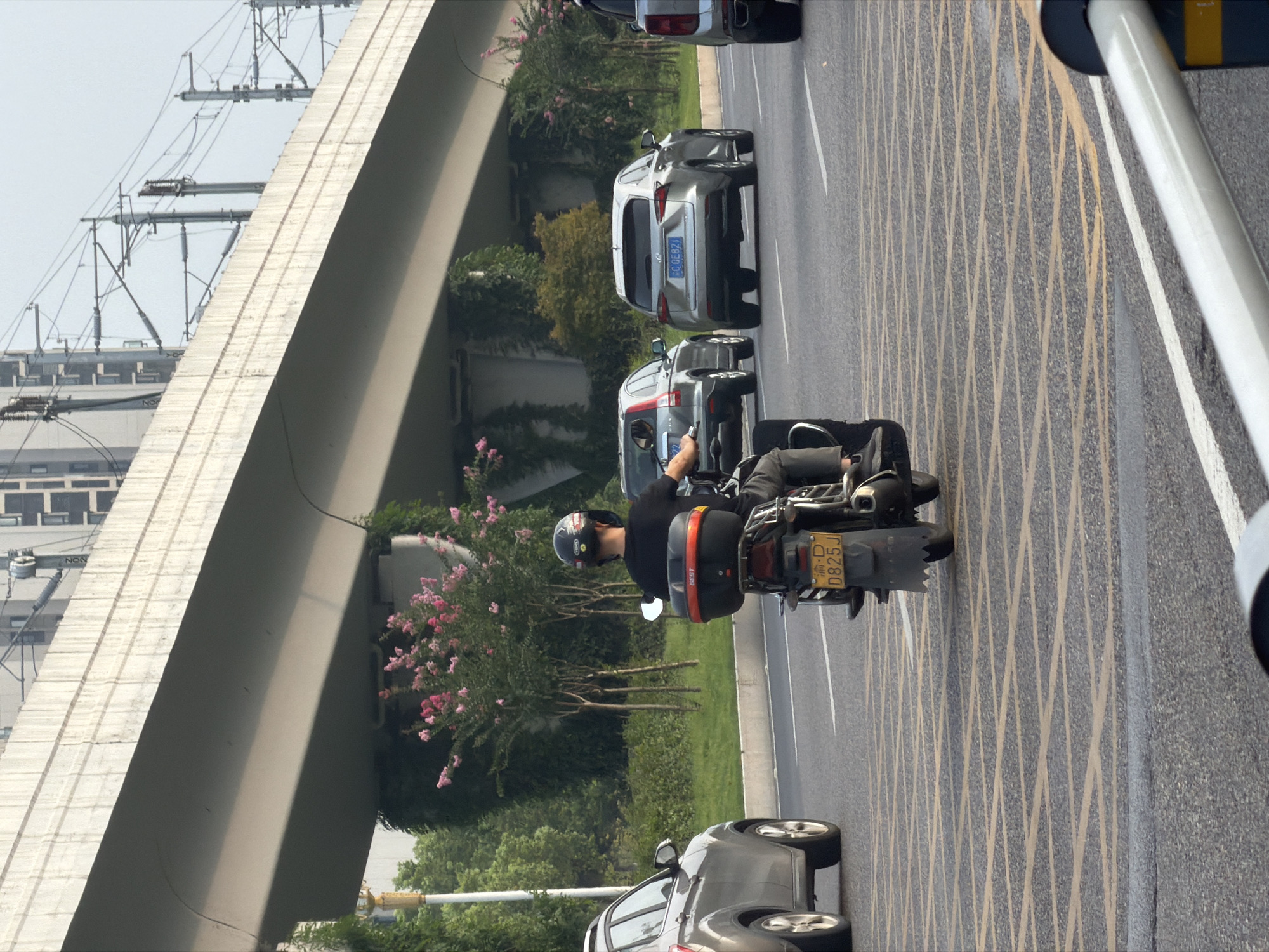 A motorcycle with a driver wearing a black helmet and black t-shirt is on a road in Jiangjin, China. The motorcycle has a black storage box on the back and a yellow license plate that reads "D825J". The road has yellow grid markings. To the left of the motorcycle is a silver SUV. In front of the motorcycle are other vehicles, including a silver Lexus SUV with a blue license plate reading "COE821". An elevated concrete overpass with train tracks runs above the road. Trees and pink flowers are visible on the side of the road. The sky is overcast.