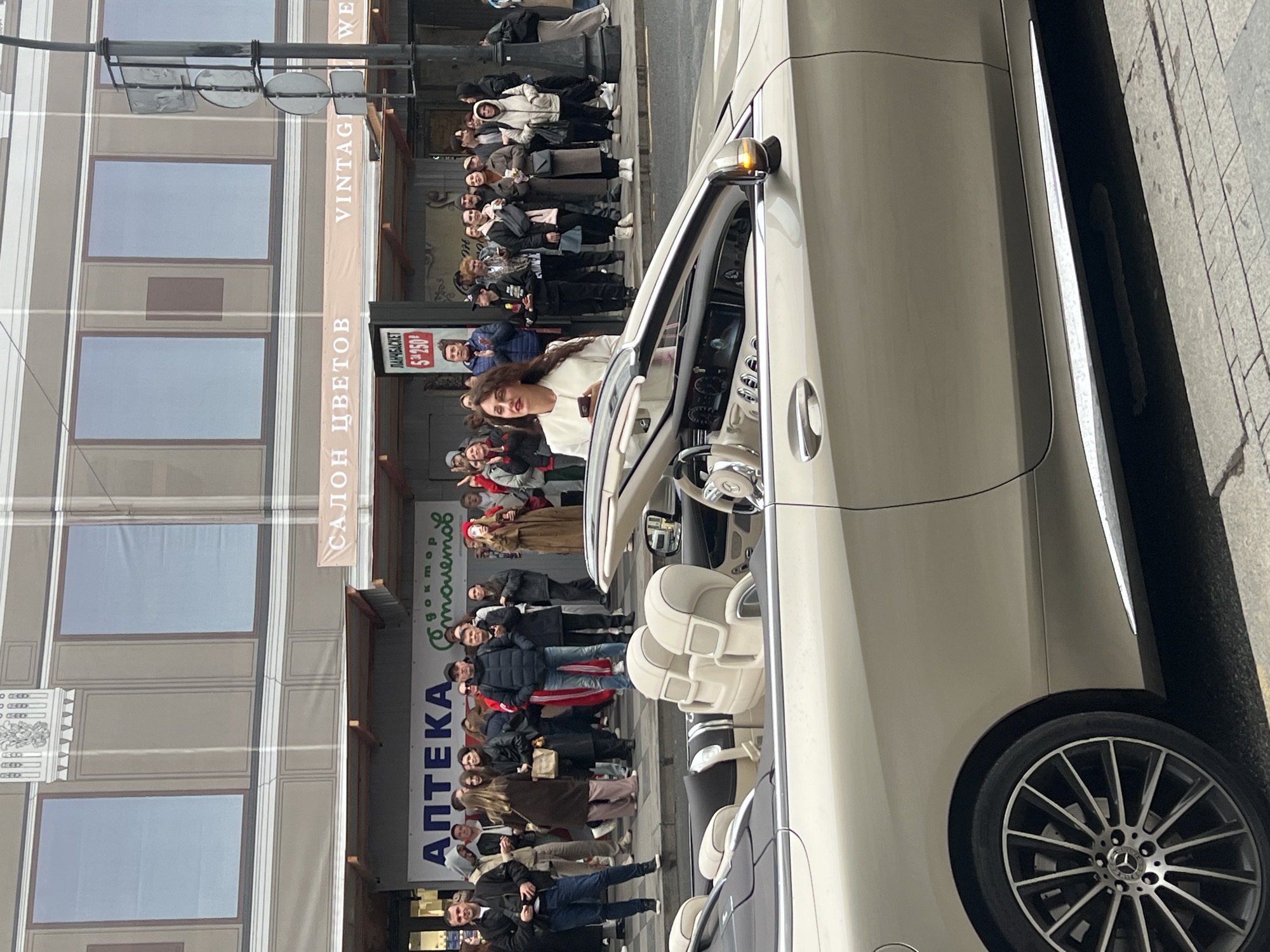 A woman is in a light-colored convertible car, with a crowd of people gathered on the sidewalk beside the car. Behind the crowd, a building has signs in Russian, including "АПТЕКА" (Pharmacy) and "САЛОН ЦВЕТОВ" (Flower Salon). A banner says "VINTAGE WED." Based on the signs being in Russian, and the city being known for this type of architecture, this is Saint Petersburg, Russia.