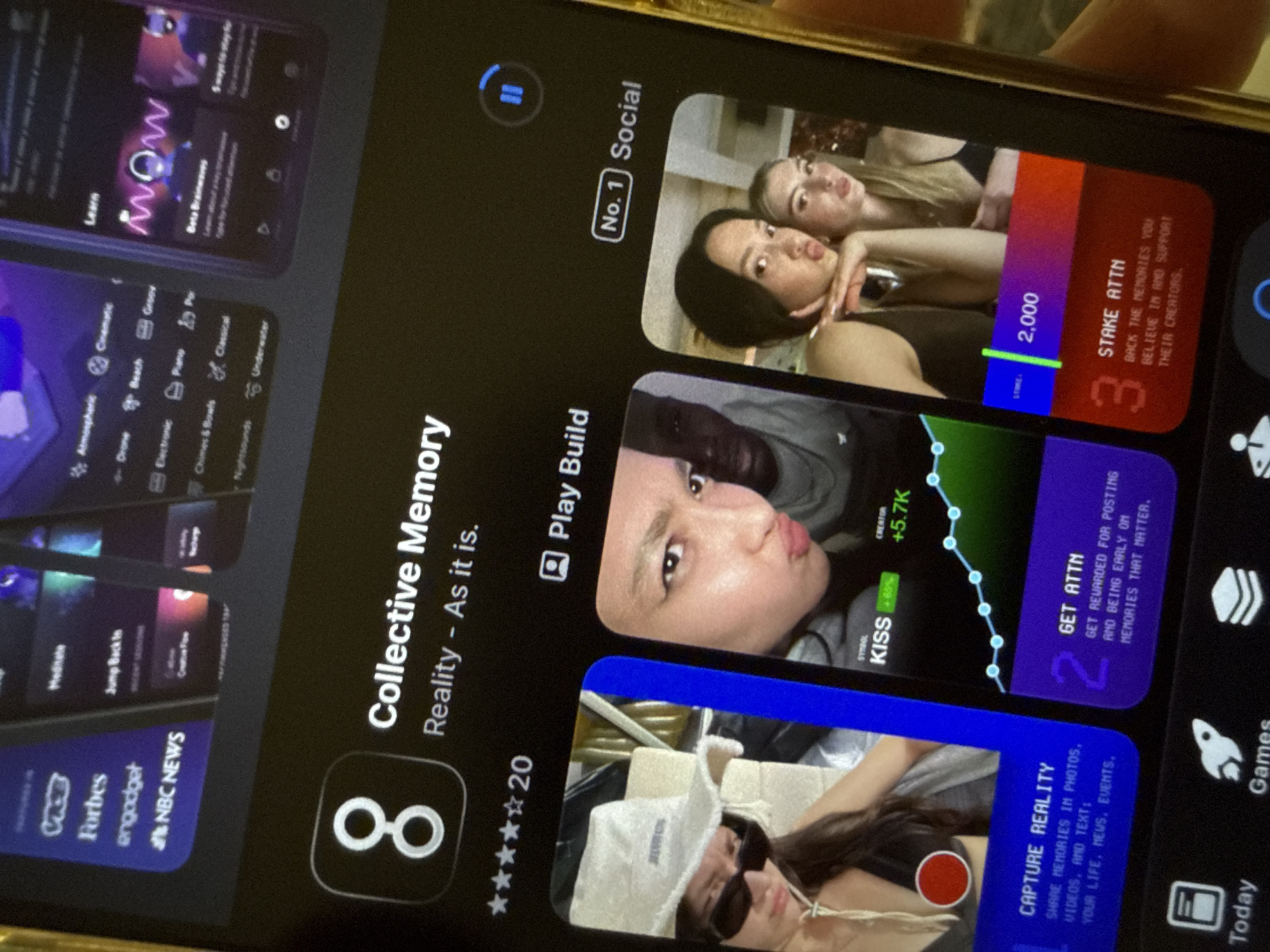 A slightly angled, close-up photograph captures a smartphone screen, likely displaying an app store interface or a specific app's page, held by a hand partially visible at the top right. The phone appears to have a golden-colored case or bezel.
On the left side of the screen, various app categories and titles are visible, including "NOW," "Learn," "Meditate," "Jump Back In," and news sources like "Forbes," "engadget," and "NBC NEWS." Categories such as "Atmospheric," "Cinematic," "Drone," "Beach," "Electronic," "Chimes & Bowls," "Nightsounds," "Underwater," "Groov," "Piano," and "Classical" are also listed.
The central focus is an app titled "Collective Memory - Reality - As it is." Its icon features two interlocking white circles on a black background. Below the title, a rating of four out of five stars is shown, followed by the number "20," possibly indicating reviews or ratings. A "Play Build" button is also present. The app is tagged as "No. 1 Social."
The right side of the screen details features of the "Collective Memory" app across three numbered sections:
* **Section 3:** Shows two young women, one with pouted lips. Text reads "STAKE ATTN" with "2,000" displayed, alongside the description: "BACK THE MEMORIES YOU BELIEVE IN AND SUPPORT THEIR CREATORS."
* **Section 2:** Features a young person with pouted lips and a graph showing "+5.7K" and "KISS +65%." The text states: "GET ATTN," "GET REWARDED FOR POSTING AND BEING EARLY ON MEMORIES THAT MATTER."
* **Section 1:** Displays a young woman wearing a hat and sunglasses, with a red recording icon overlaid. Text advises: "CAPTURE REALITY," "SHARE MEMORIES IN PHOTOS, VIDEOS, AND TEXT: YOUR LIFE. NEWS. EVENTS."
At the bottom right of the screen, parts of a navigation bar with icons for "Today" and "Games" are visible. The image is bright with screen reflections.