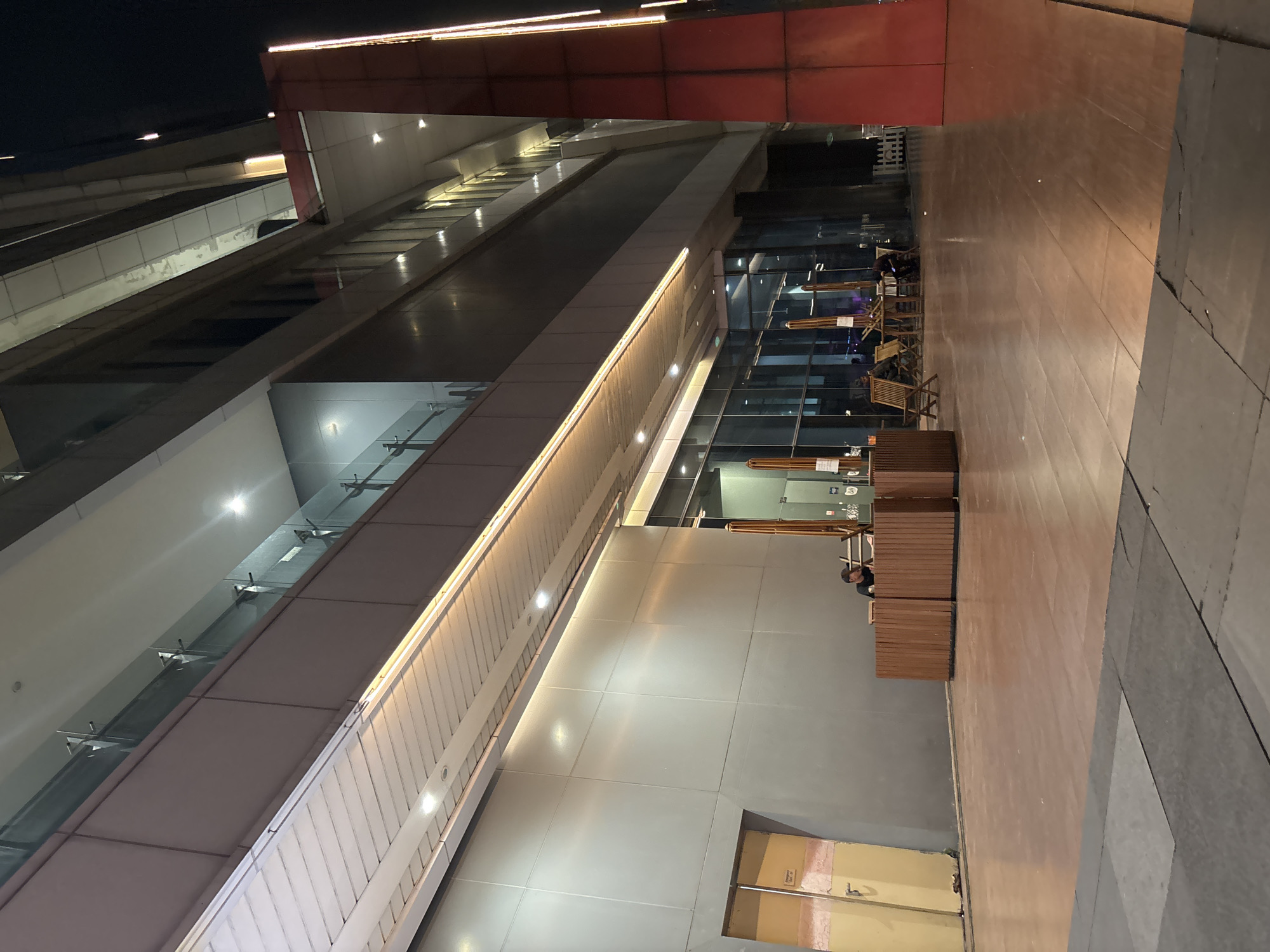 A nighttime exterior view of a modern building in North Jakarta, Indonesia, shows a covered walkway with wooden flooring. A man in a dark shirt and baseball cap is standing behind a slatted wooden counter. To the right, several tables and chairs with umbrellas are set up. The building features large glass windows and is illuminated by warm, artificial lights. A tall, red pillar stands prominently on the right side of the frame, also lit by vertical lights. The foreground consists of textured pavement.