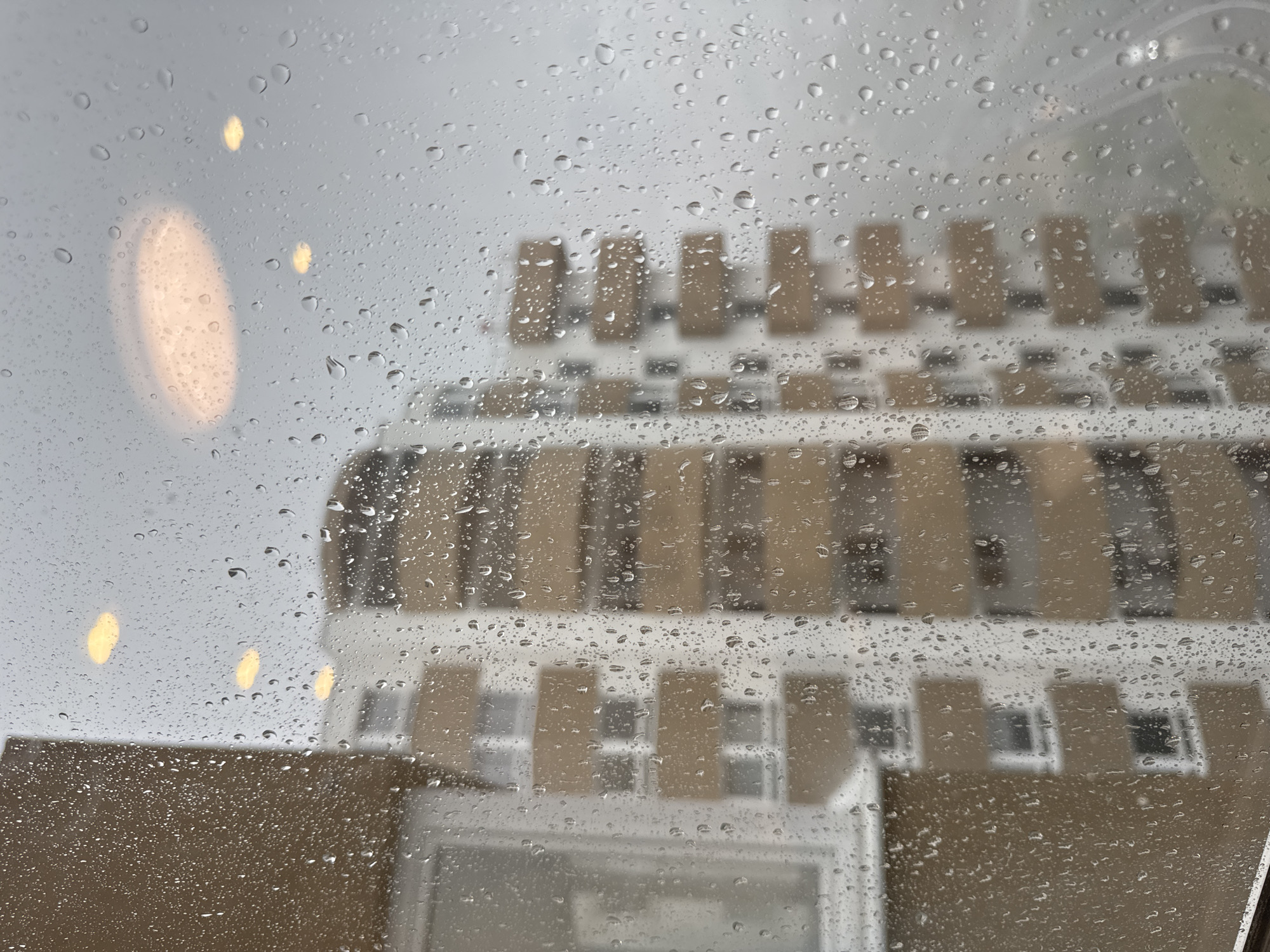 The image depicts a view through a rain-streaked window, looking out at apartment buildings in Sochi, Russia. The window is covered in numerous water droplets, obscuring the view and creating a blurred effect. In the foreground, to the left, a portion of a building's exterior wall, with a textured brown finish, is visible. A window with a white frame is also present on this side.
The main subjects of the image are two multi-story apartment buildings in the background. These buildings feature a modern architectural style with repeating balconies. The balconies appear to have a light brown or beige color, contrasting with the white facade of the buildings. The buildings are seen through the rain-covered glass, making their details softened and slightly distorted.
Above the buildings, in the sky, are several out-of-focus circular lights. These appear to be ceiling lights or lamps, possibly from within the building from which the photo is taken, or from streetlights. The overall atmosphere is grey and overcast, consistent with rainy weather. No people or animals are visible in the image. The scene conveys a sense of being indoors on a rainy day, observing the urban landscape.