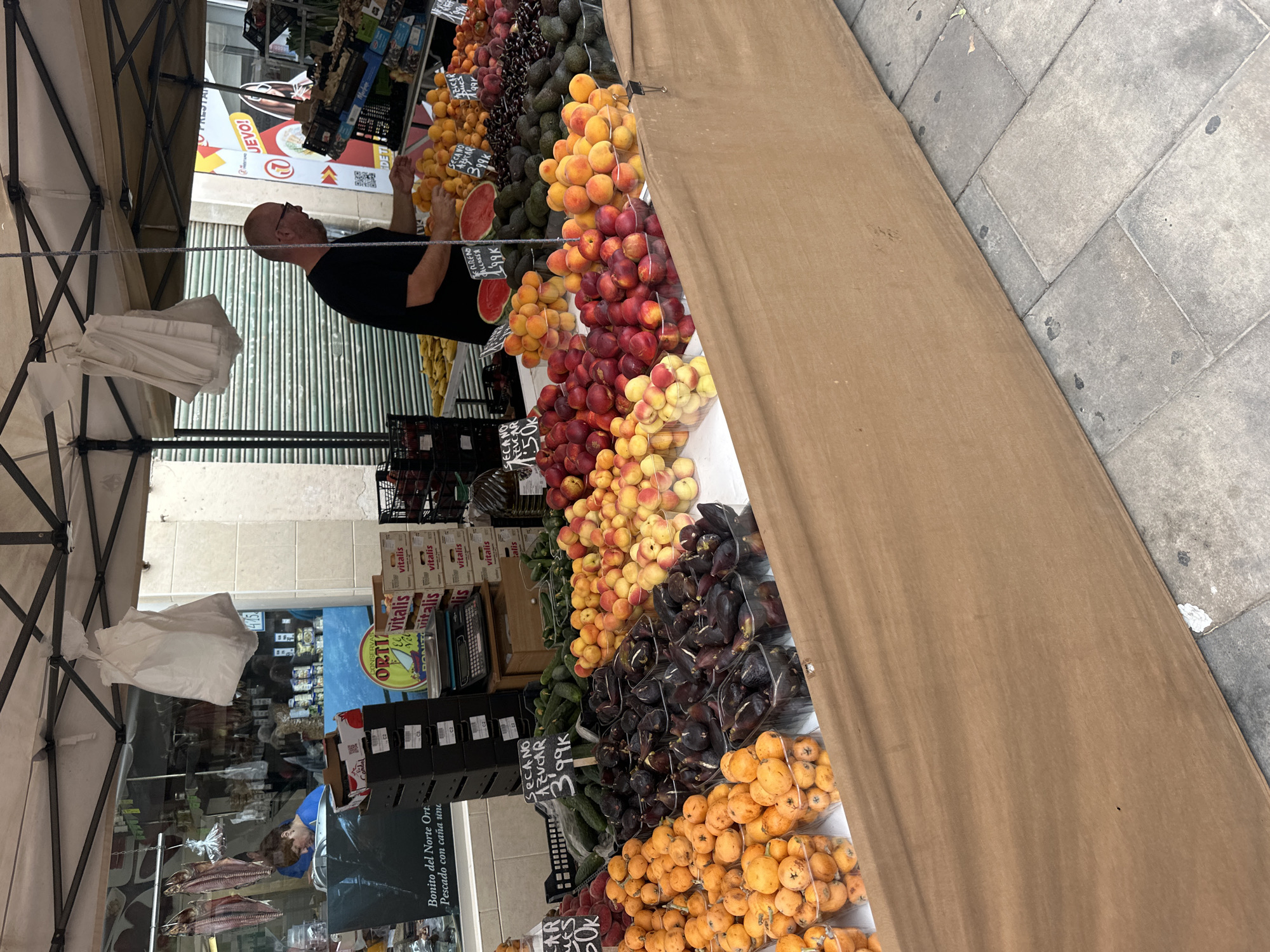 An outdoor market stall, brightly lit during the daytime, showcases a vibrant array of fresh fruits and vegetables. The stall is covered by a light-colored canopy, with its framework visible at the top and left of the image.
In the center, a bald man wearing a black t-shirt is visible in profile, standing behind the stall. He appears to be a vendor, with his hands near a sliced watermelon, possibly arranging or serving produce. His expression is not clearly discernible. Further back and to the left, another person's head and shoulder are partially visible behind a counter inside what appears to be a shop.
The market display features numerous items, including piles of ripe peaches and nectarines, darker purple plums or figs, bright orange loquats, green avocados, bananas, and cucumbers. Many fruits are neatly arranged in clear plastic bags or containers. Various price signs are interspersed among the produce. The ground in front of the stall is paved with grey and tan flagstones.
Behind the stall, a shopfront is visible through an open entrance, with shelves displaying products. Inside the shop, several smoked or dried fish hang from a rack. Boxes labeled "Vitalis" are stacked, and a sign for "CONSERVAS ORTIN" is present.
Visible text includes: "SECANO AZÚCAR 399K" (likely pricing for a type of dry/sugar-free produce, repeated), "SECANO AZUCAR 1.50k", "TERRENO ALLOSSA 1.99K", "Bonito del Norte Ort. Pescado con caña une" (referring to line-caught Northern Bonito tuna), "CONSERVAS ORTIN", "Vitalis", and partially visible words like "JEVO!", "RESTA...", and "DETI". The language suggests a Spanish-speaking region.