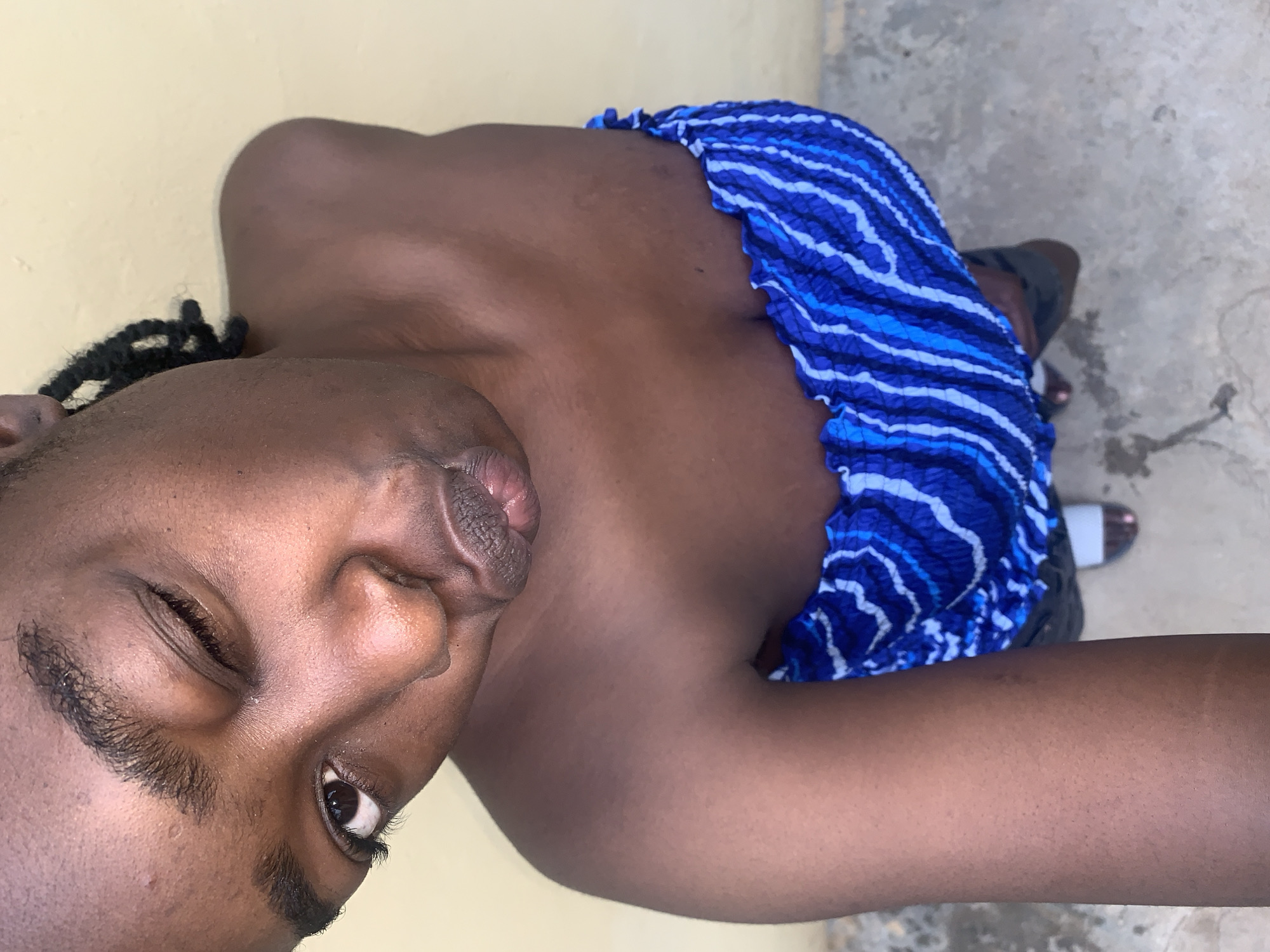 A high-angle shot captures a person with dark skin looking upwards directly at the camera. The individual winks with their left eye, which is closed, while their right eye is open and focused. Their lips are pursed forward in a "duck face" or kissing gesture. Dark hair styled in braids or dreadlocks is visible on the right side of their head. They are wearing a strapless garment, either a top or a dress, which has a ruched elasticated neckline and features a distinct wavy or irregular horizontal stripe pattern in various shades of blue and white. Their bare shoulders and upper chest are visible. The background consists of a plain, light yellow-tan wall behind the upper body, transitioning to a grey, textured concrete ground surface in the lower portion of the image. Partially visible dark open-toed sandals with white accents are on the person's feet. The scene takes place in Rafin Par, Nigeria.