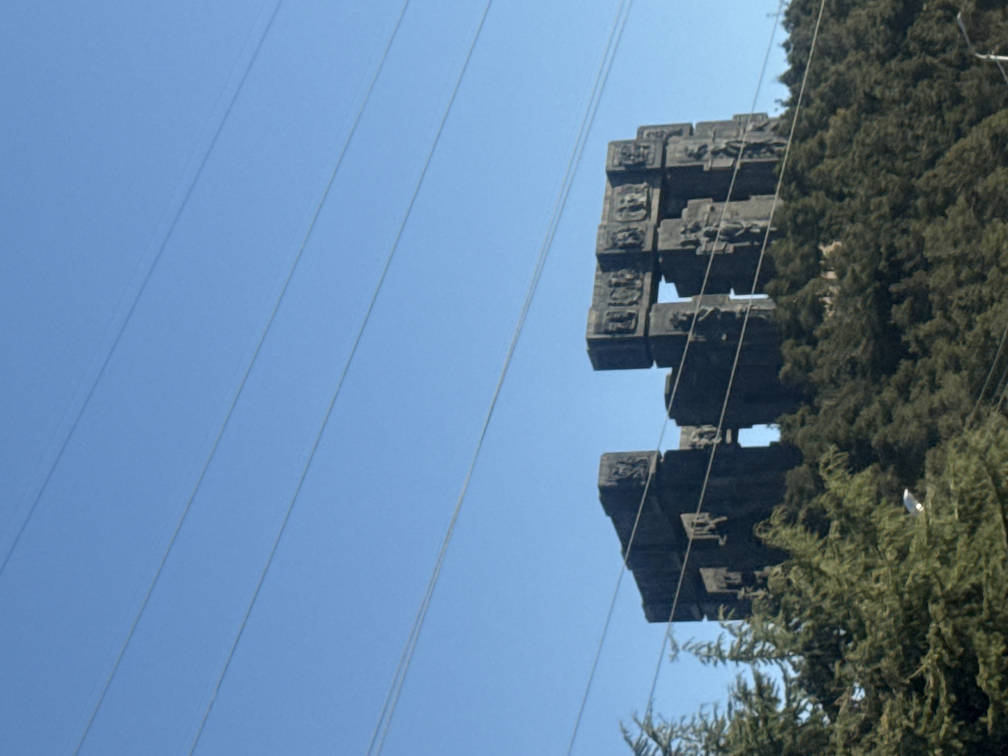The image, taken from a low angle, features a section of the Chronicles of Georgia monument in Tbilisi. The monument, a large structure made of concrete, is partially obscured by trees. The monument features relief carvings on its surface. Above the monument, several power lines are visible against a clear blue sky. The trees adjacent to the monument are evergreen, with varying shades of green foliage. The sky is cloudless.