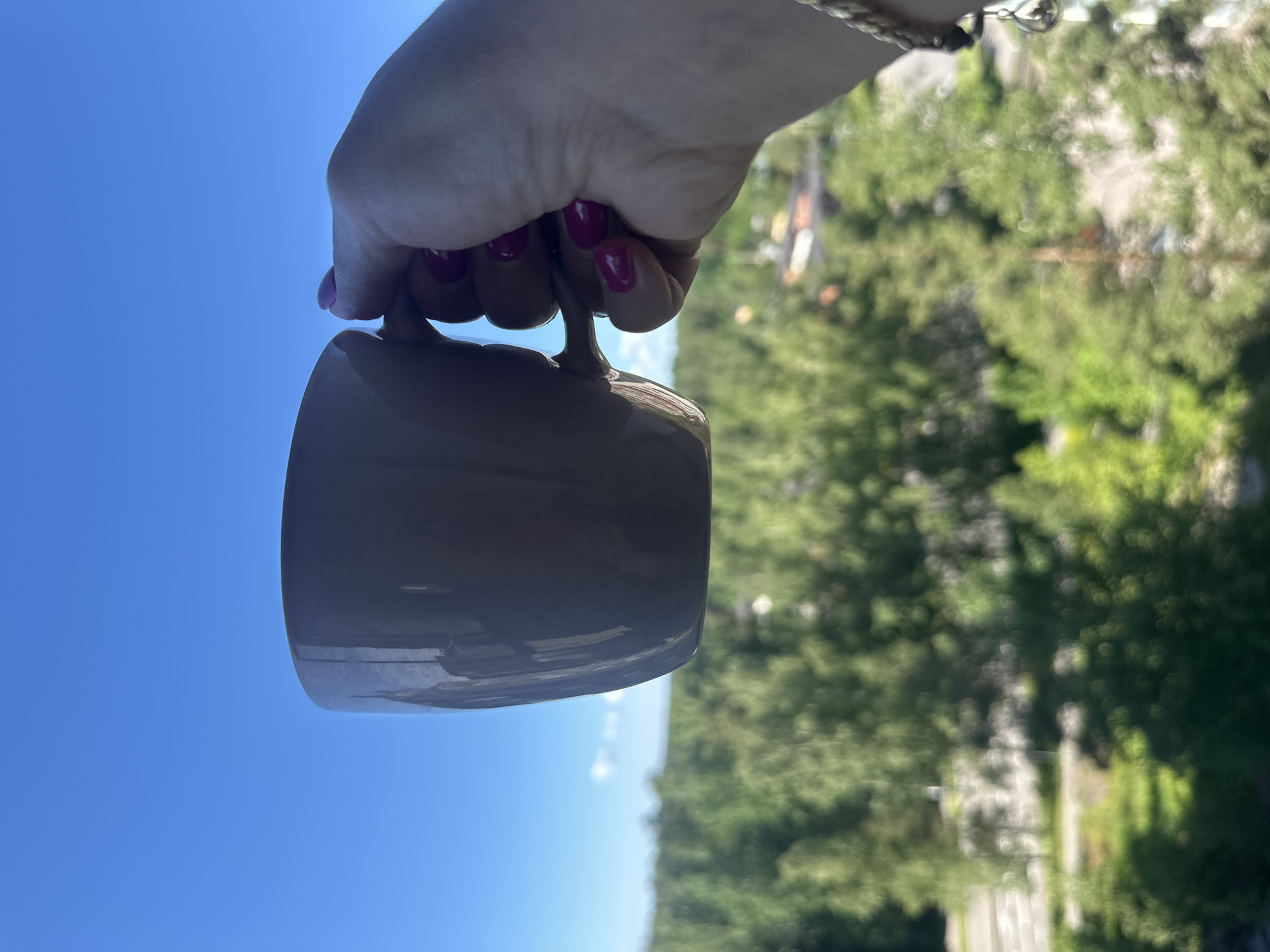A hand with bright pink nail polish holds a beige mug against a bright blue sky. The mug is positioned in the foreground and partially obstructs the view of a lush, green forest in the background. Hints of buildings are visible through the trees. The lighting suggests it is daytime.