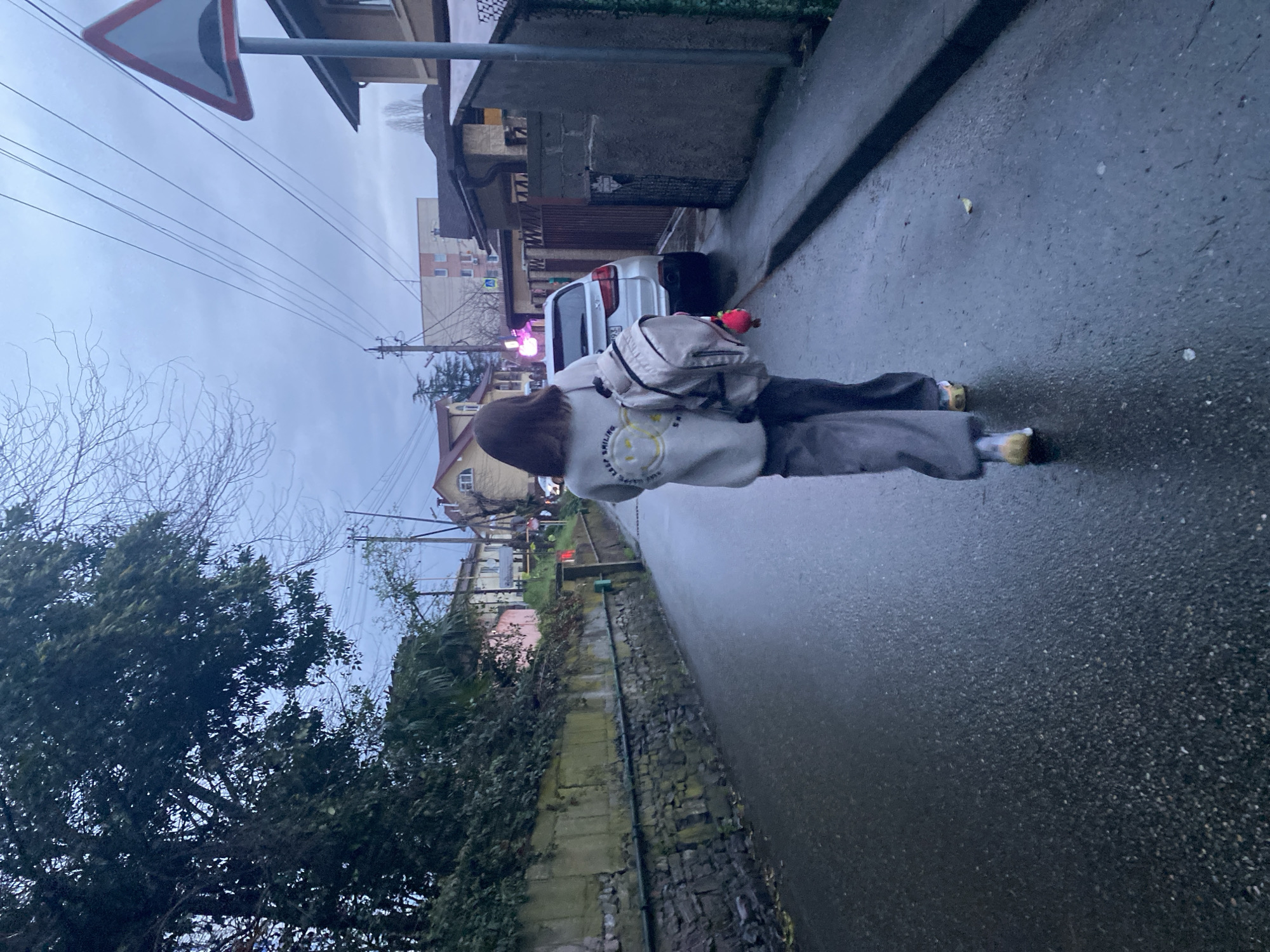 A young woman is walking on a wet street in Sochi, Russia. She is wearing a gray sweatshirt with a smiley face on the back, gray pants, and yellow shoes. She has a backpack on her back and is holding a red object in her hand. A white car is parked on the side of the road. The sky is overcast. There are buildings, trees, and power lines in the background. A road sign is visible in the top left corner of the image.