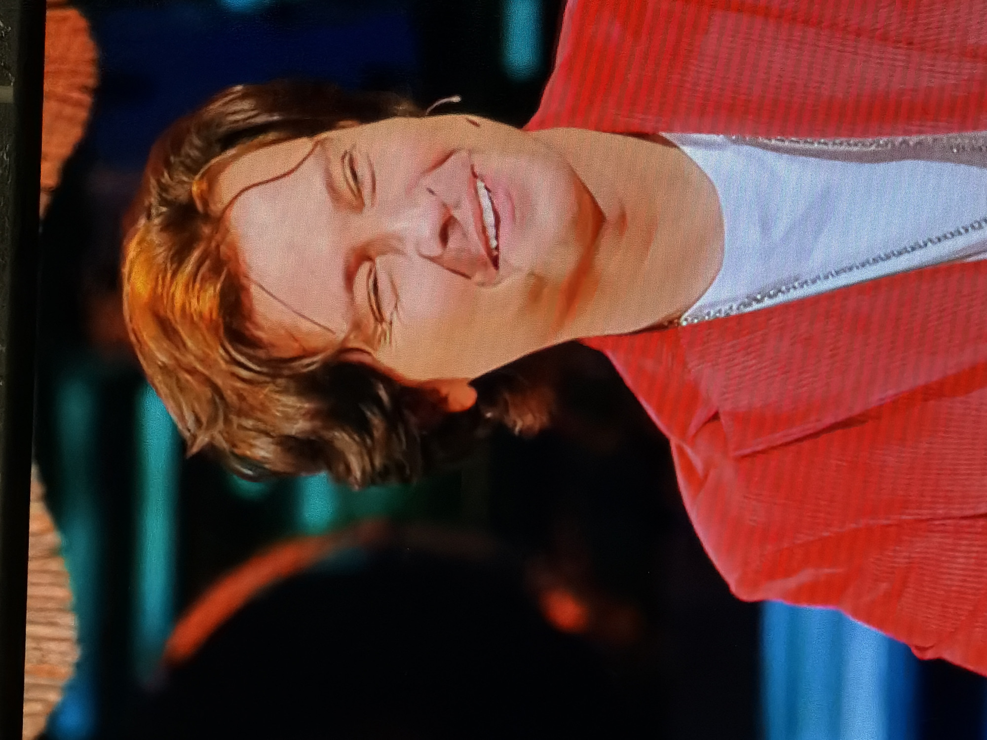 The image depicts a fair-skinned individual, likely a woman, with short brown hair styled with highlights, smiling with her eyes partially closed. She is wearing a white shirt with a chain necklace underneath a red corduroy jacket. The background is blurry but appears to be an indoor setting with blue and orange lighting. There is no information about the location being in Moscow, Russia.