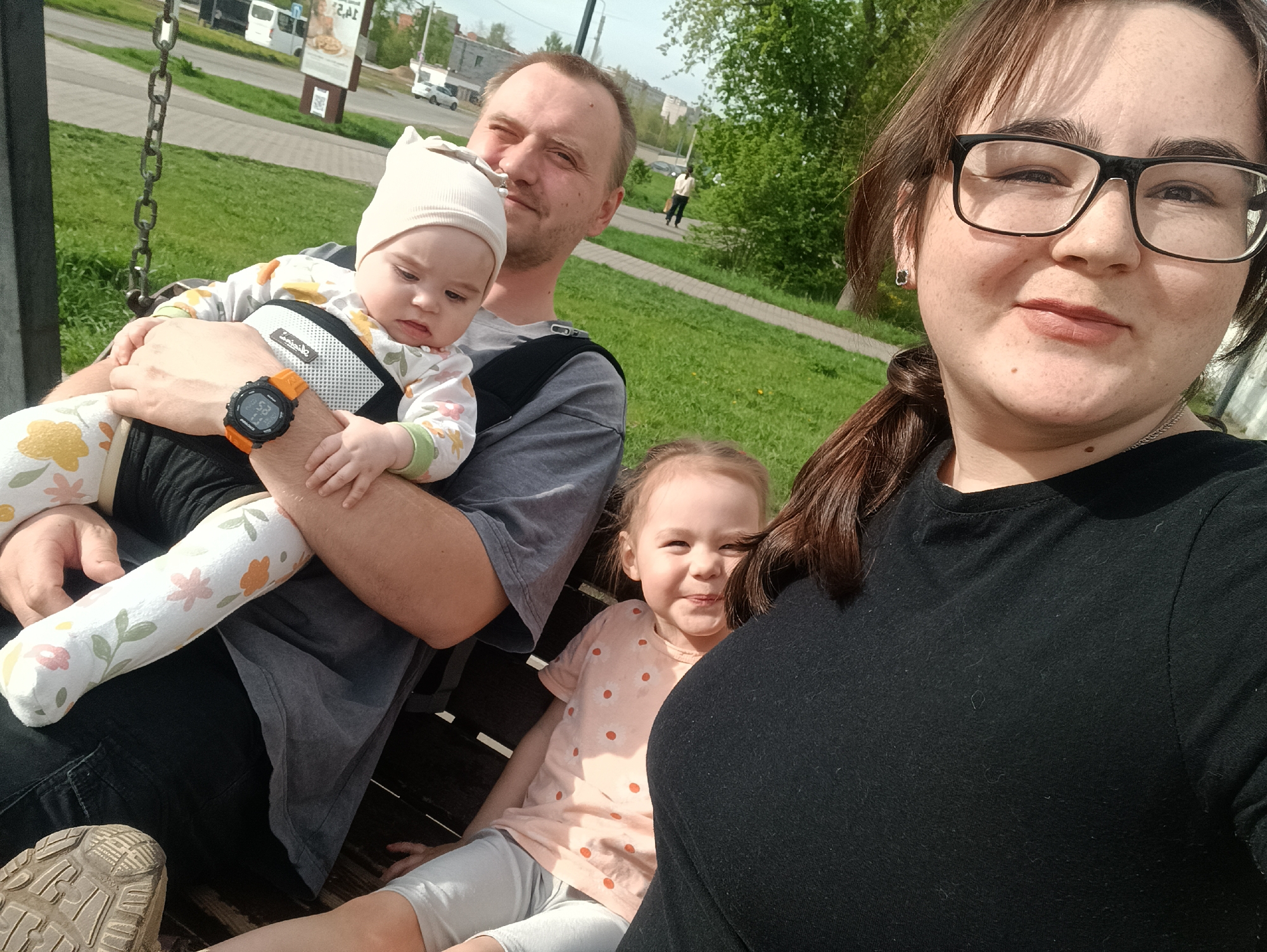 A group of four people, presumably a family, are captured in a photograph taken in what appears to be a public park in the city of Chaikovsky, Russia. The group consists of a man, a woman, and two young children. The location is identifiable by a billboard displaying a Russian Cyrillic advertisement. The father is holding an infant wearing a white floral outfit and a white hat, and wearing an orange wristwatch. The older daughter is seated on the bench, smiling. The woman, who is wearing glasses and a black shirt, appears to be taking the photo with a mobile device. The environment is well-lit, suggesting daytime, and the background includes greenery, a sidewalk, and a street with vehicles. The family is seated on a swing-like bench with chains, indicating a park or recreational area. The man is wearing a gray T-shirt. The infant is secured in a baby carrier. The woman has brown hair, pulled back in a ponytail. The older child is wearing a light pink shirt with an orange flower pattern and grey pants. The photograph appears to be a casual snapshot.