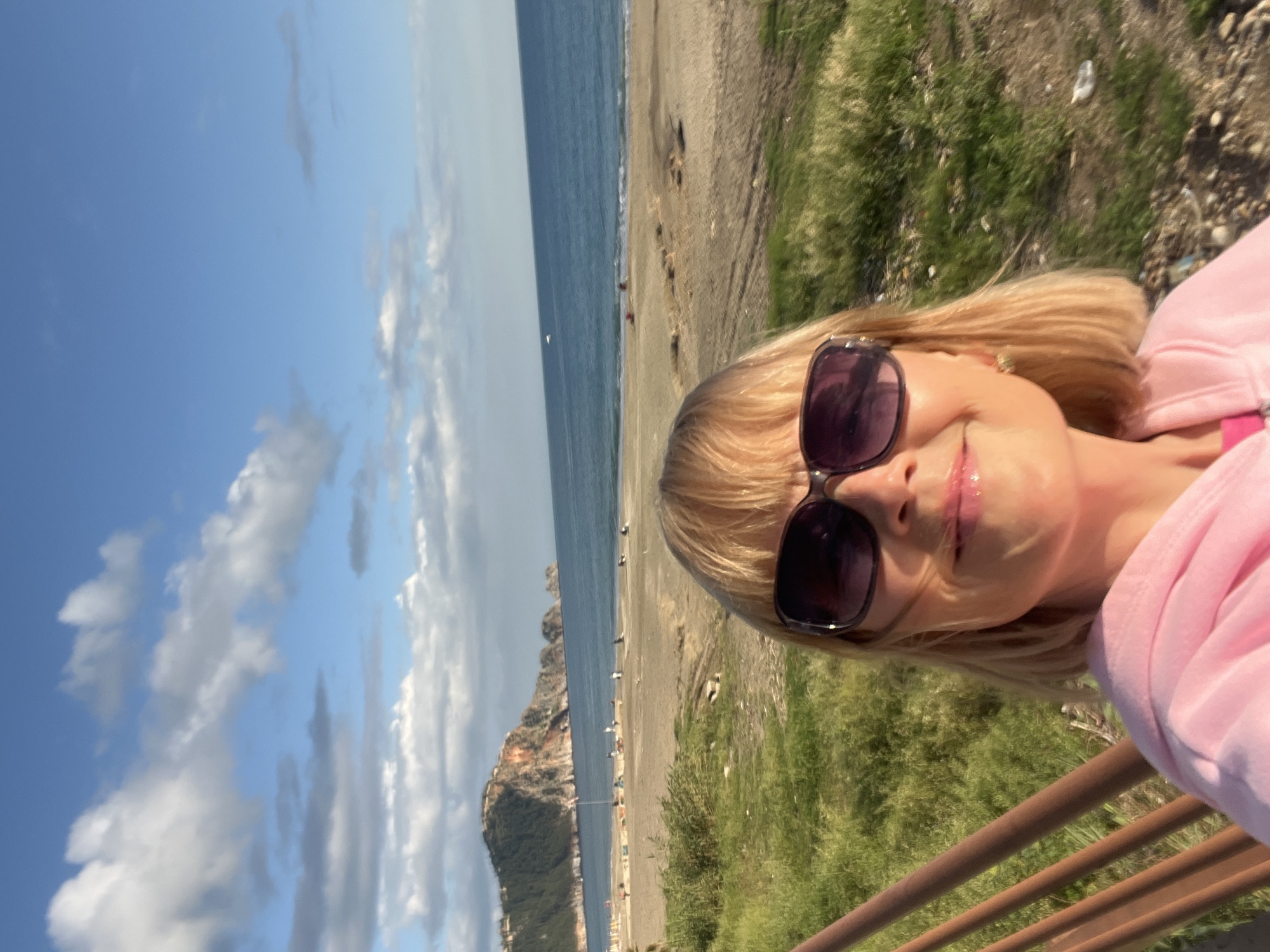 A woman is taking a selfie. She is wearing sunglasses and a pink top. The background features a beach, the sea, a rocky headland, and a blue sky with clouds. The location is Alanya, Turkey. There is a brown railing in the foreground.