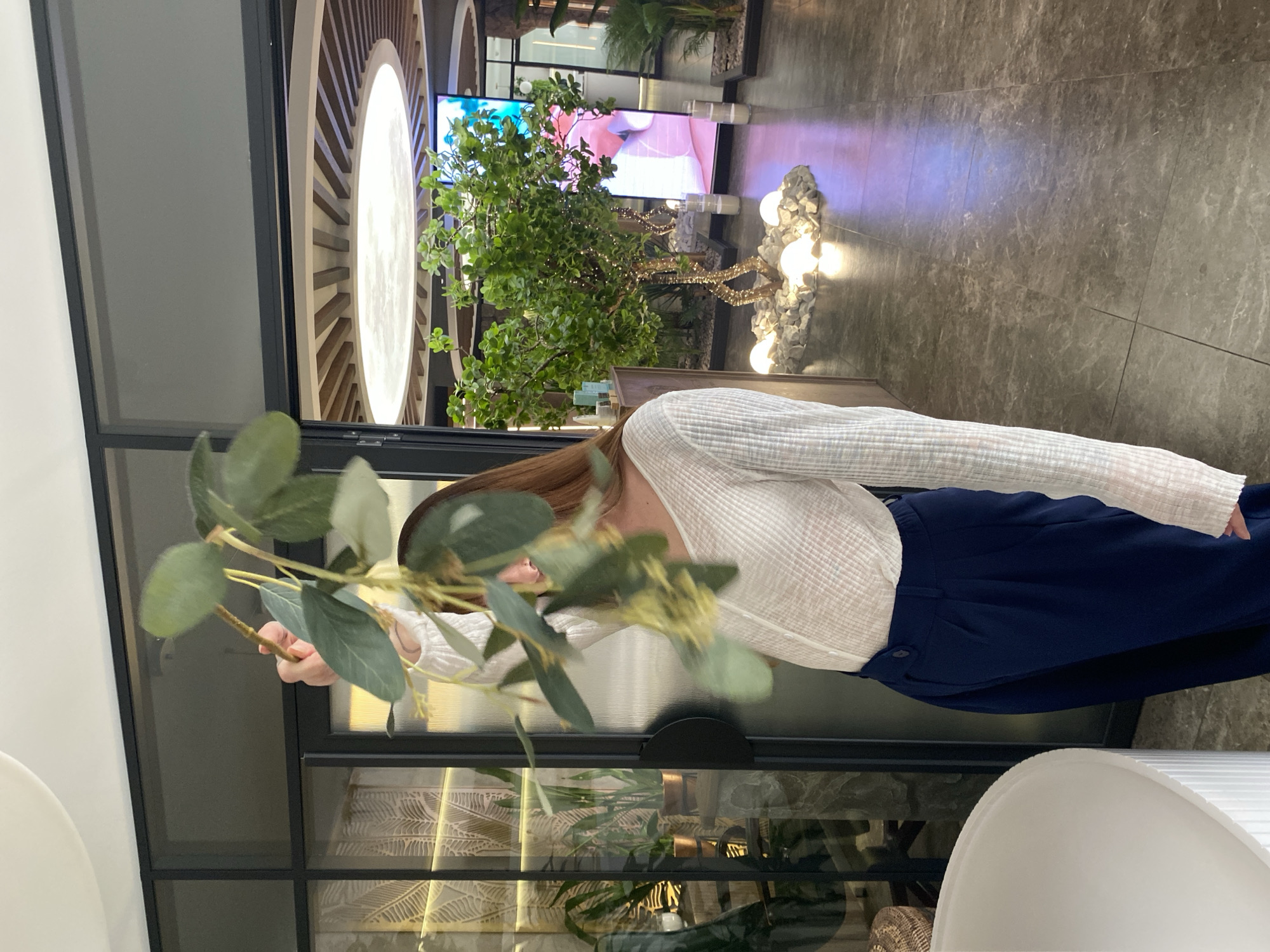 A medium shot captures a woman partially obscured by a leafy branch. She is seen from the waist up, wearing a white, long-sleeved textured top and dark blue pants. She appears to be holding the branch.
The background features a modern interior. Elements include a black-framed window or room divider and decoratively patterned walls. In the distance, there's a lighted tree-like structure, possibly an artificial display with light bulbs, along with a screen or monitor displaying bright imagery.
There is no indication in the image that it was taken in Votkinsk, Russia.