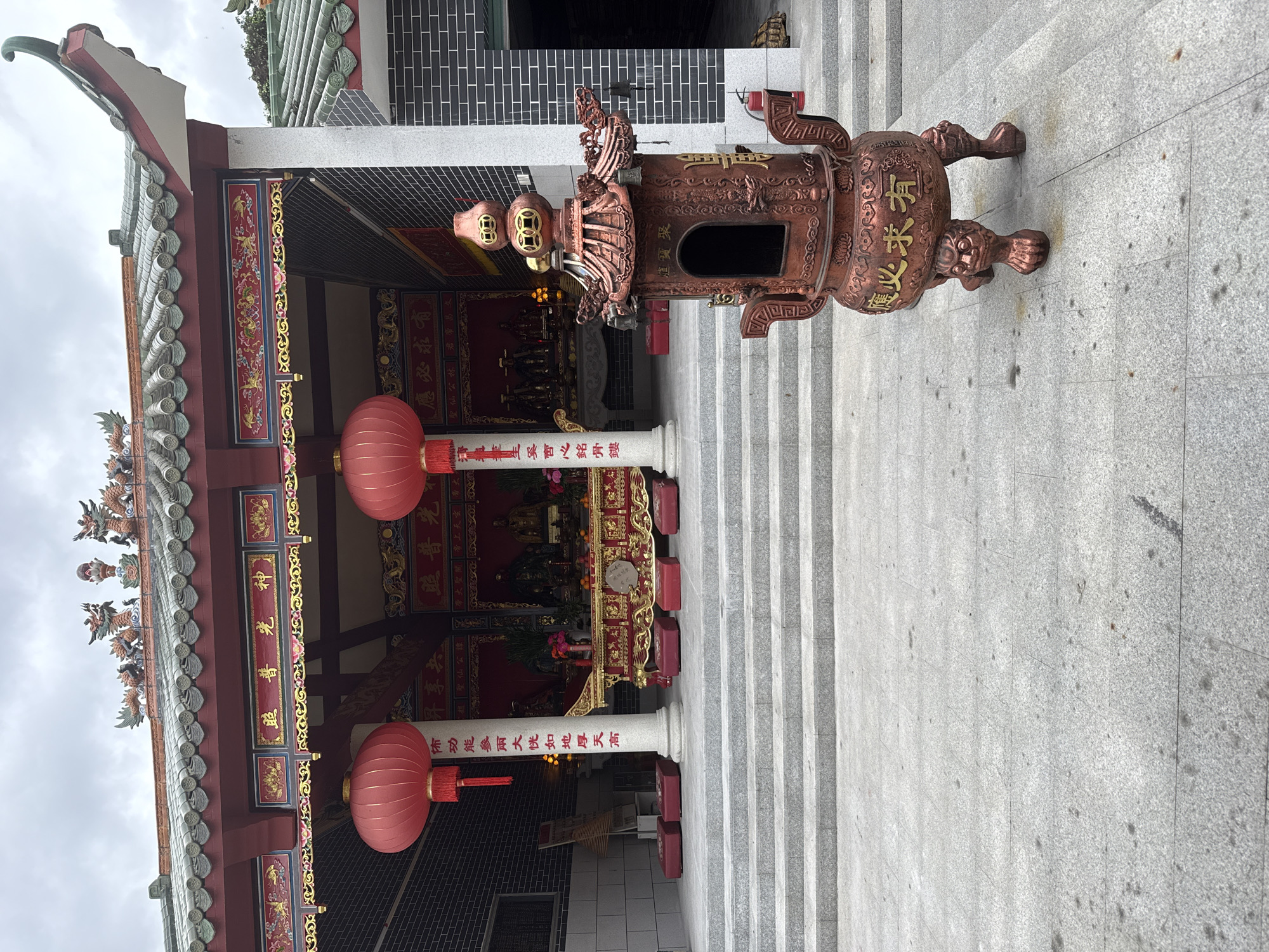 The image depicts a scene in front of a traditional Chinese temple, possibly located in Huizhou, China. The temple has a red and green tiled roof adorned with dragon figures. Red lanterns hang from the entrance, and steps lead up to the temple's altar, where several statues are visible. In the foreground, a large, ornate bronze incense burner stands on a stone-tiled courtyard. The sky is overcast, suggesting a cloudy day. There are no people visible in the image. The overall scene is one of religious architecture and traditional cultural elements.