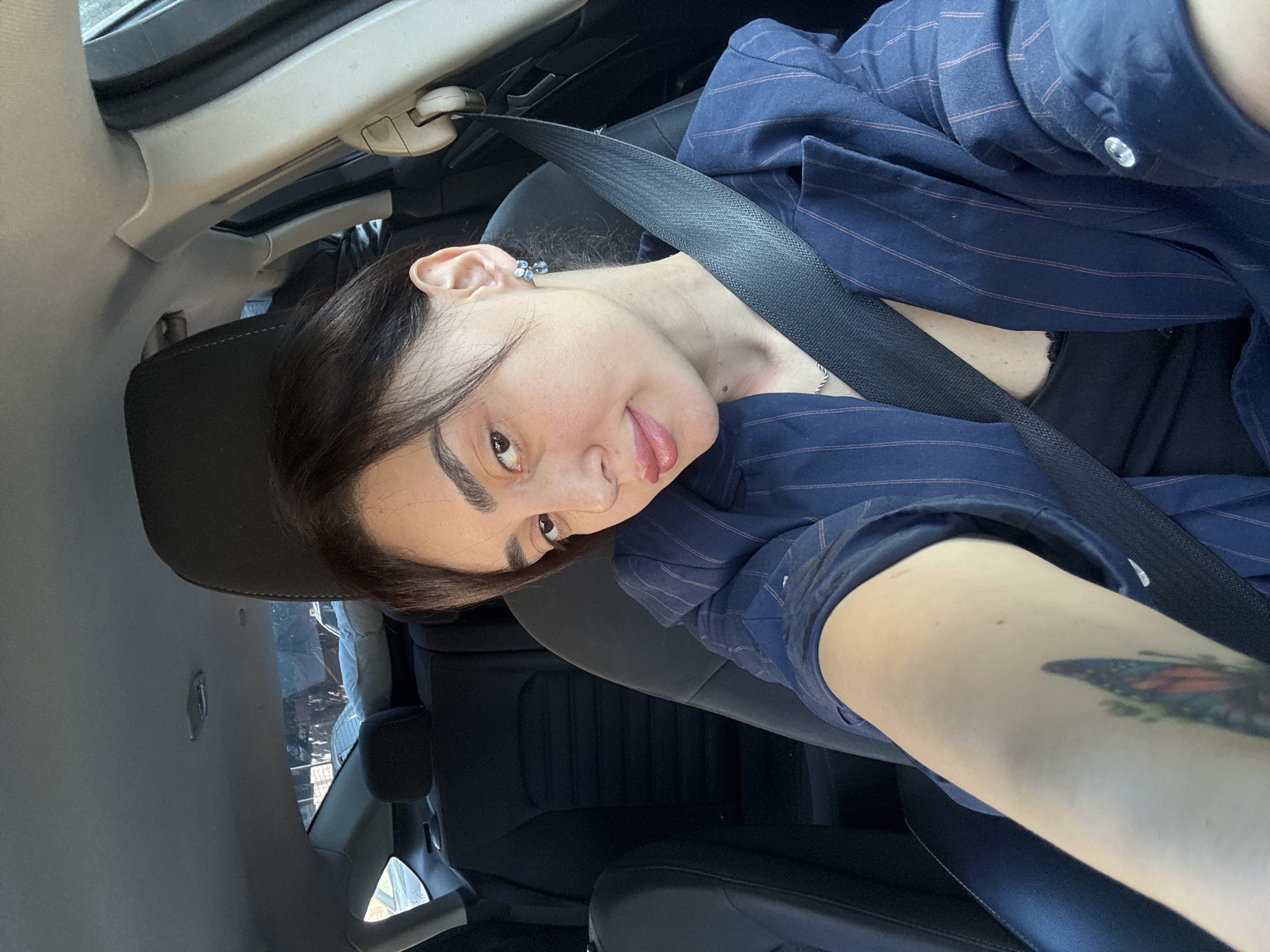A woman is photographed from a low angle inside a car. She wears a blue pinstriped shirt and has a black seatbelt on. She is sticking her tongue out. A tattoo is visible on her arm. The car's interior is visible. The location appears to be Tbilisi, Georgia, judging by the exterior view through the window.